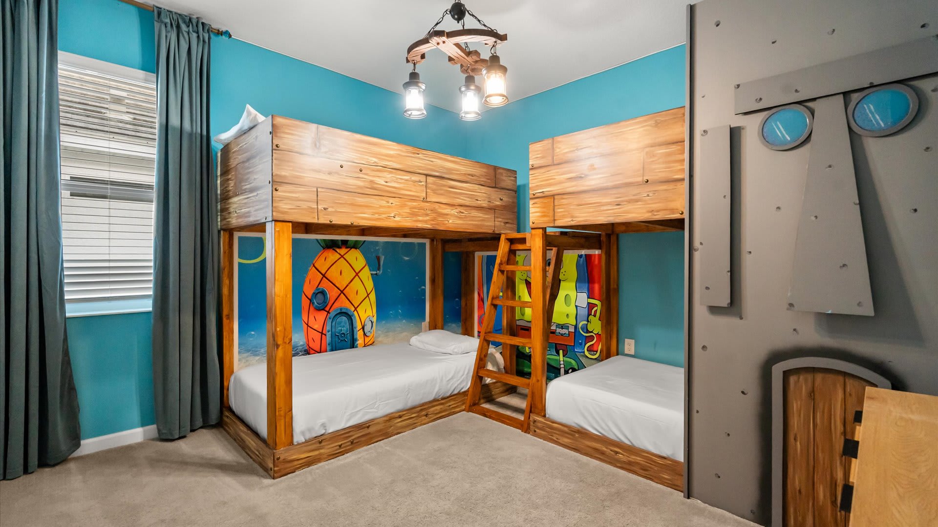 A vibrant Spongebob Squarepants themed bedroom featuring two custom wooden bunk beds with ladders. The walls are painted bright blue, and a large mural of Spongebob and his pineapple house is visible.