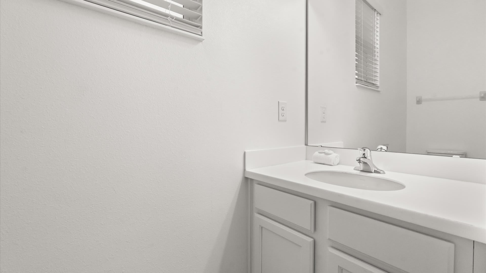 A simple, bright white bathroom features a single vanity with an oval sink, a mirror, and a window with blinds, complemented by a small white towel on the countertop.