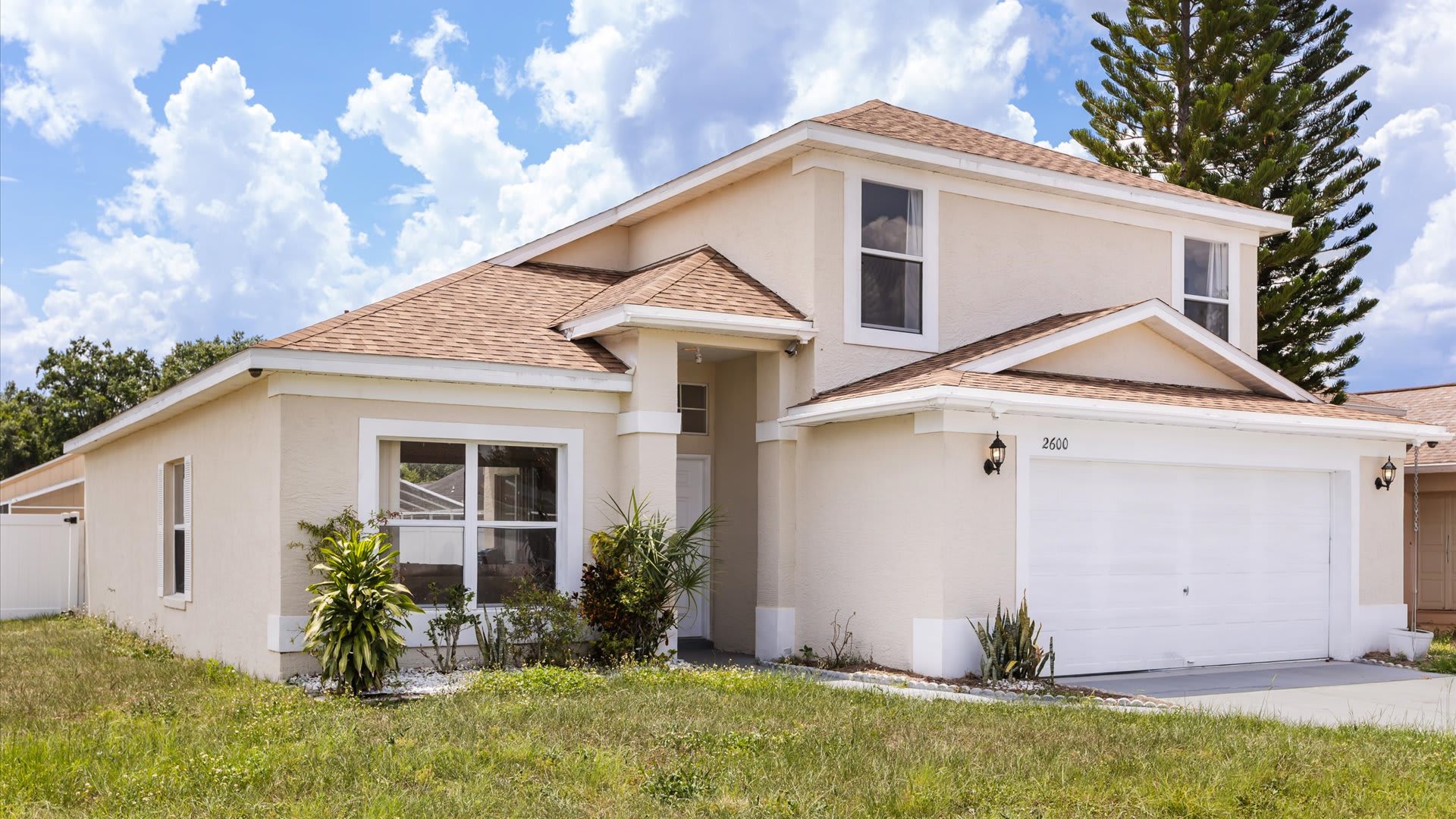 A slightly angled view of the front of a two-story, light-colored stucco house with a brown shingle roof, showing the two-car garage, front entrance, and a small landscaped garden area.