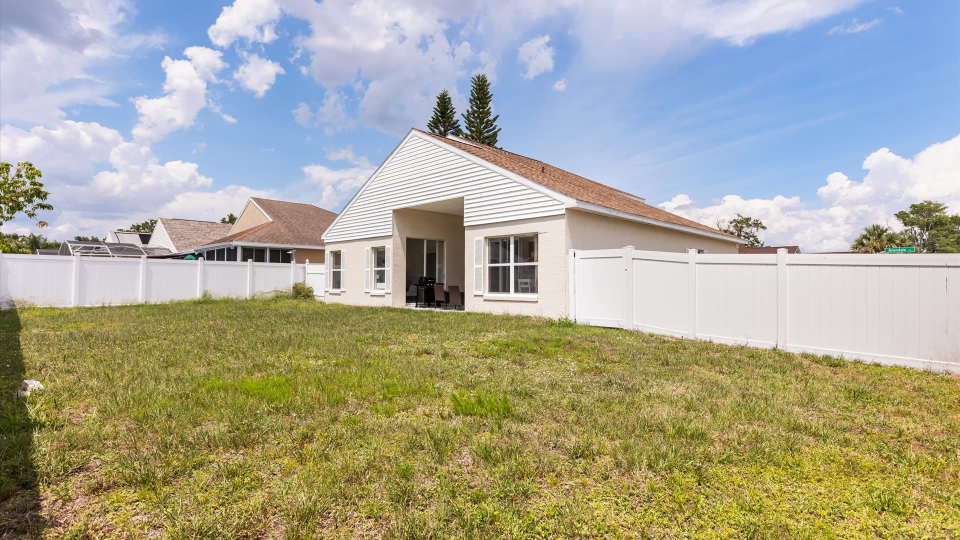 The back of a single-story house with light-colored stucco walls and a brown shingle roof, featuring a covered patio area and multiple windows, surrounded by a large grassy backyard and a white privacy fence.