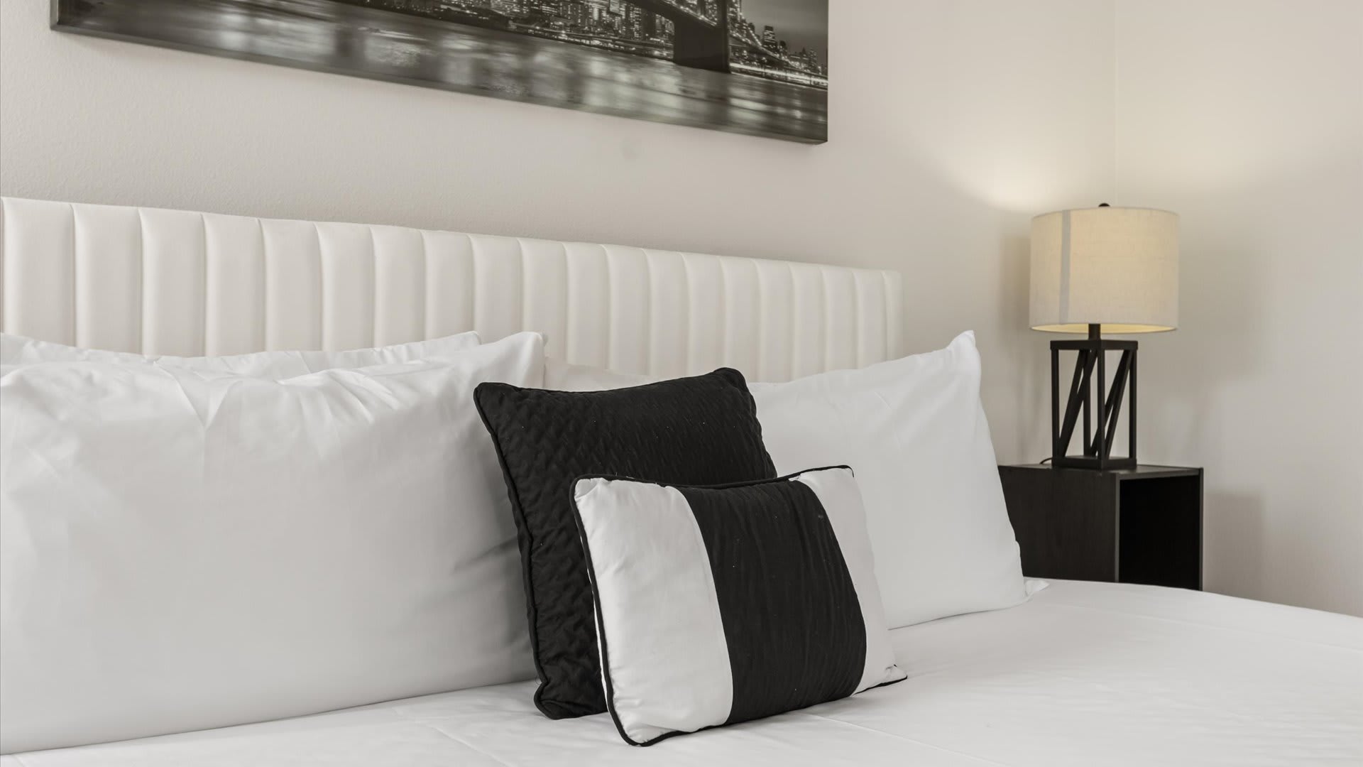 Close-up of a bed with a white upholstered headboard and white bedding, accented with black and white decorative pillows. A modern bedside lamp is visible on a dark nightstand.