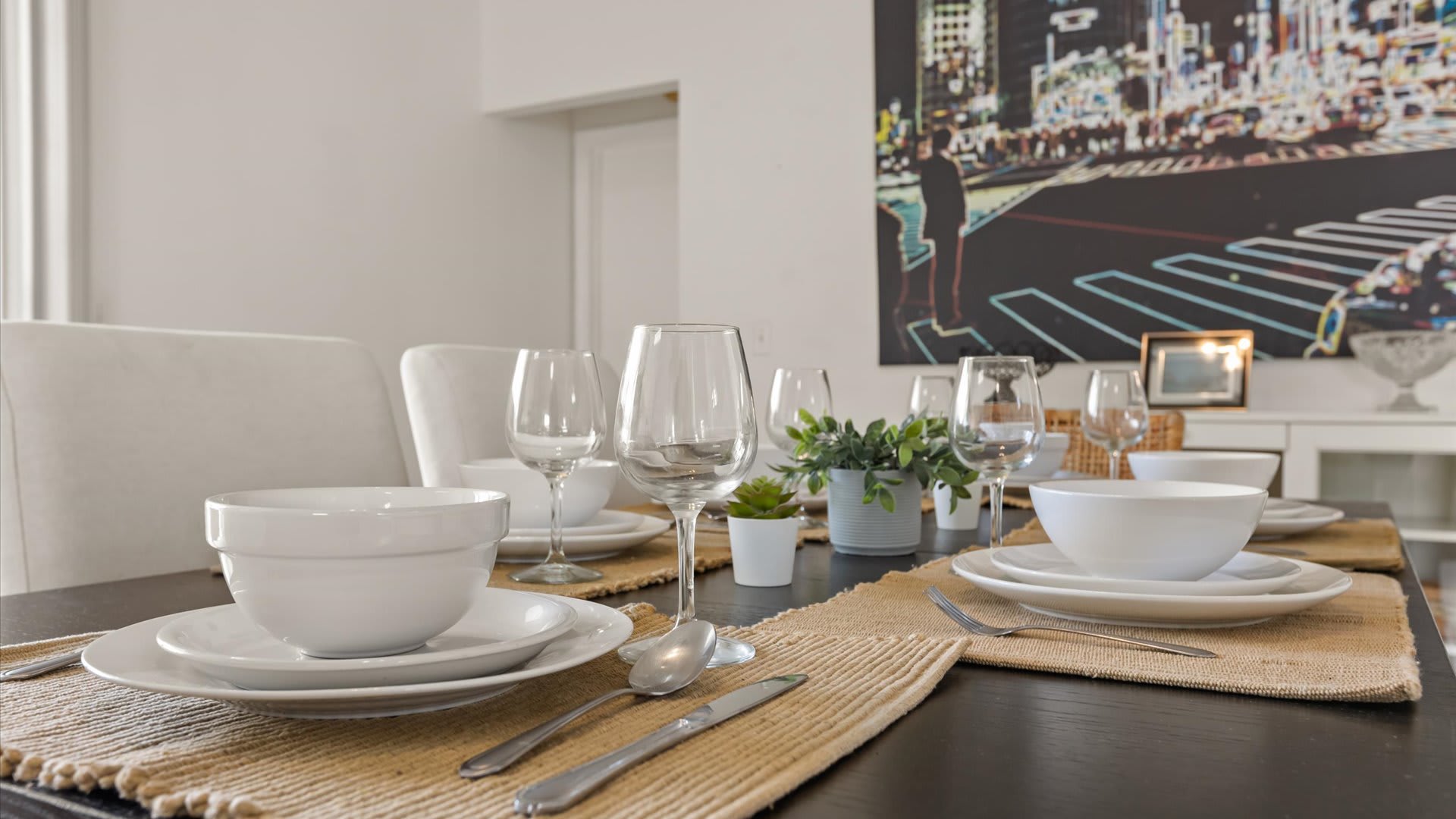 Close-up of the dining table set with white dishes, wine glasses, and placemats, featuring a small potted plant centerpiece. A large cityscape artwork is visible in the background.