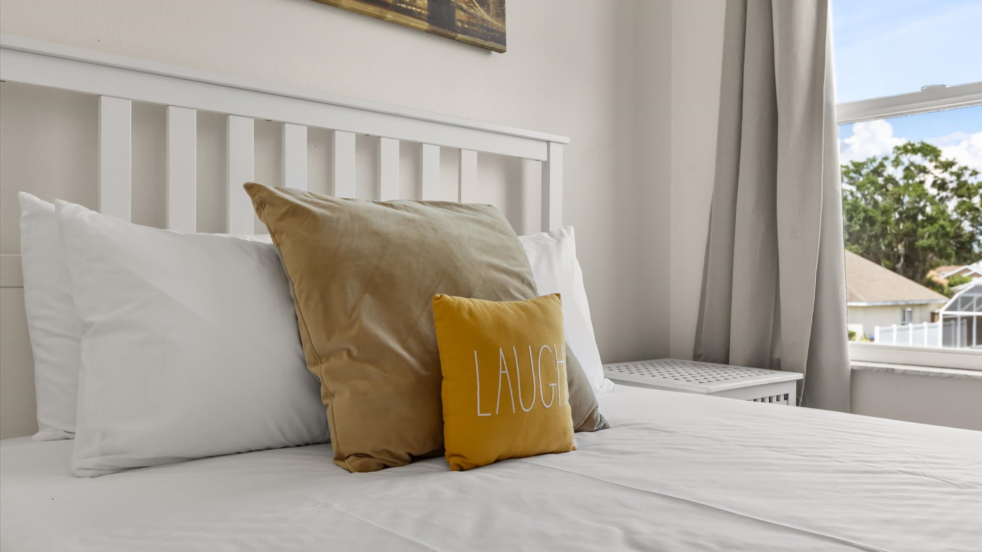 A close-up of a white wooden bed with crisp white bedding, accented by a large gold pillow and a smaller yellow "LAUGH" pillow. A white nightstand is visible to the right.