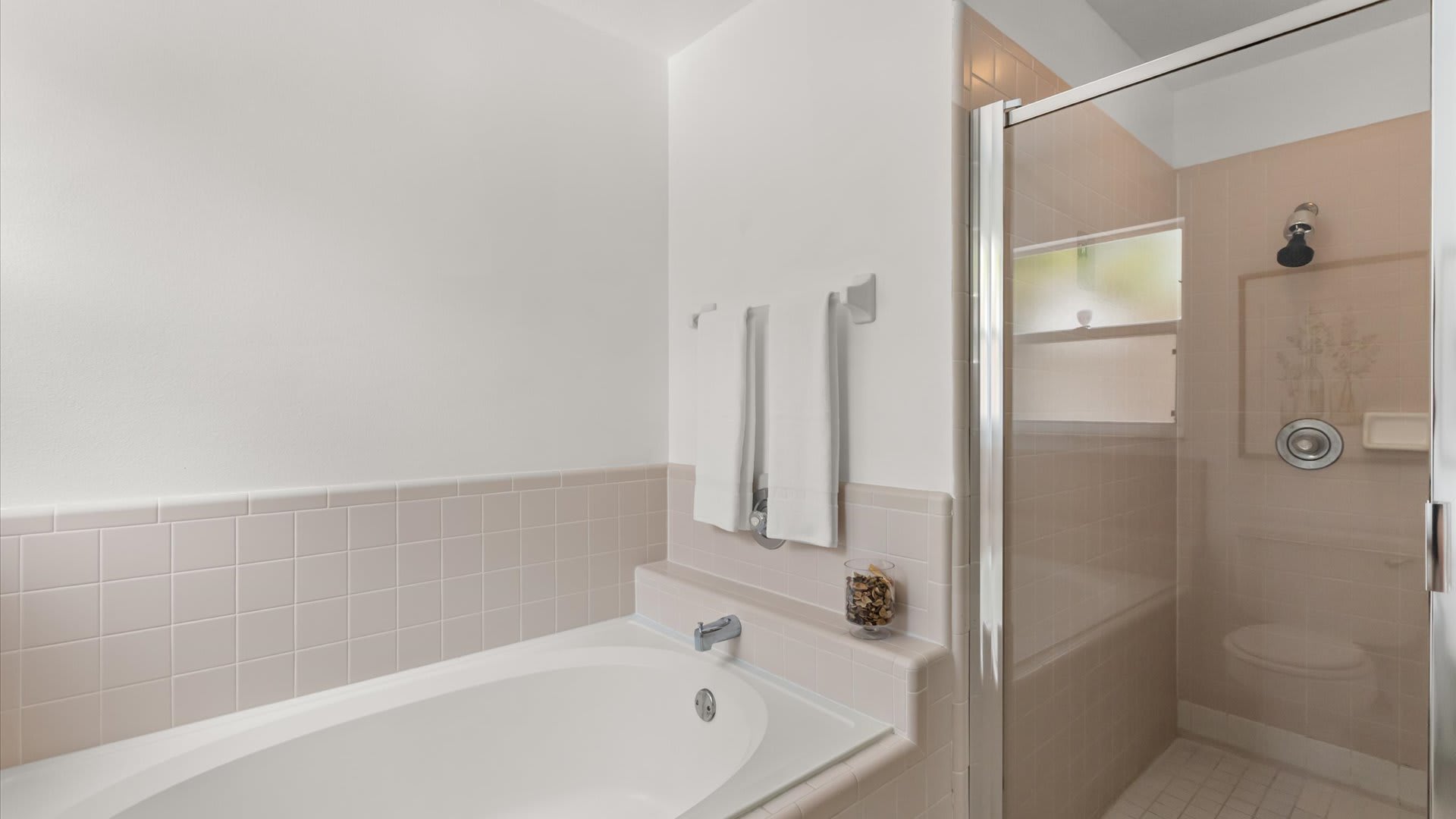 A close-up view of the built-in bathtub and glass-enclosed shower in a bathroom, featuring light pink tiled walls. White towels hang on a rack next to the tub.