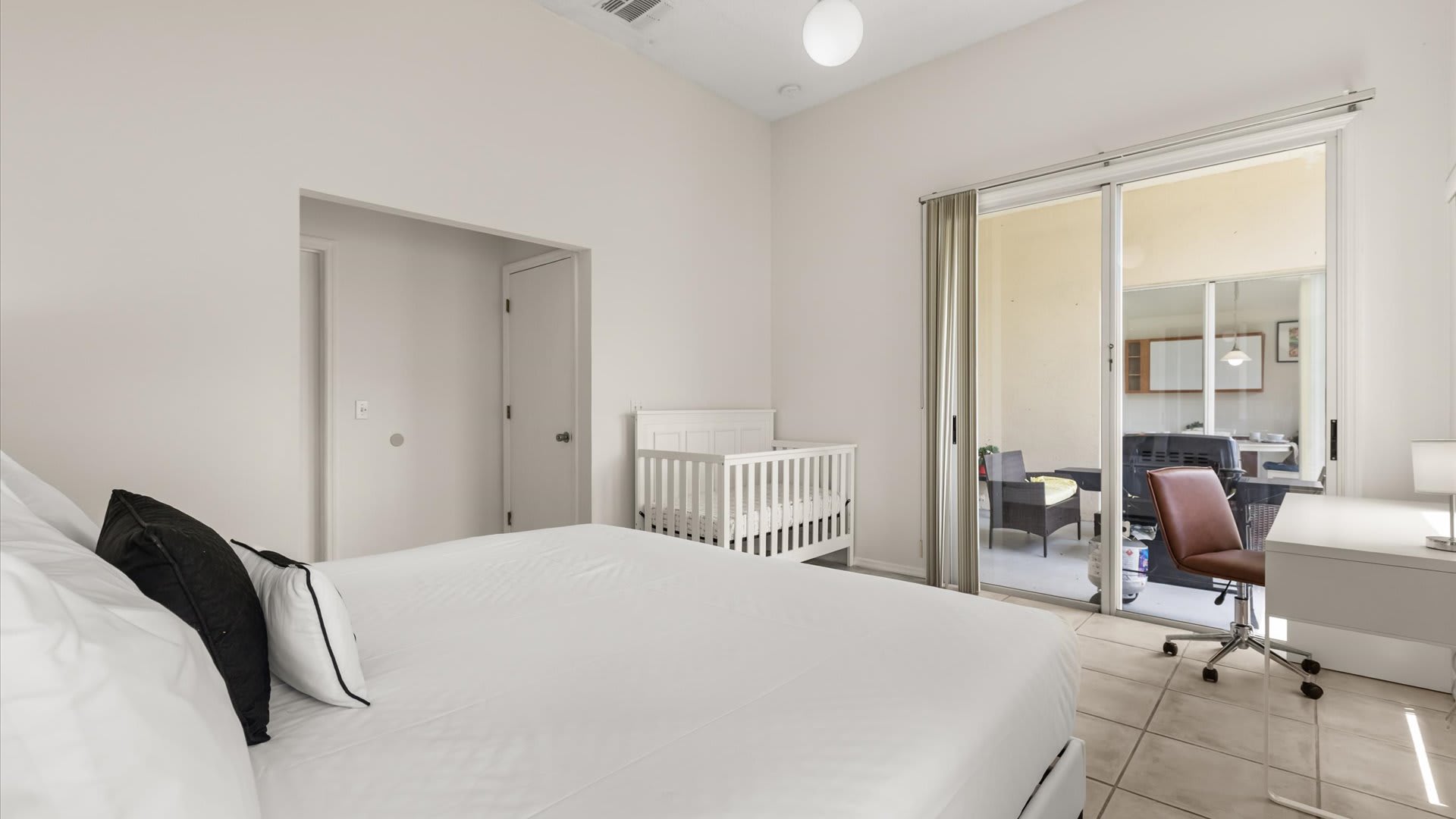 Bright bedroom with a large bed, a white crib, and a desk with an office chair, featuring sliding glass doors that open to an outdoor patio. The room has white walls and tile flooring.