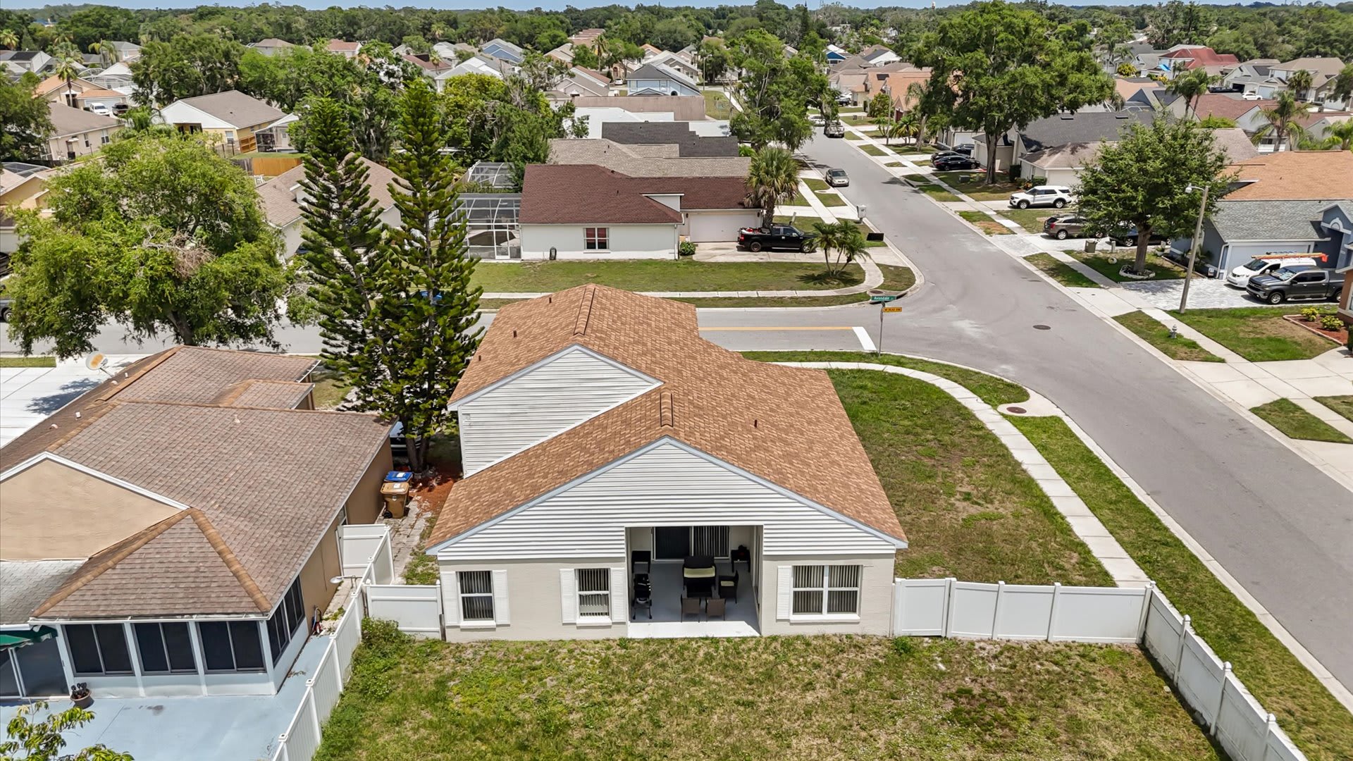 An aerial view of a light-colored house with a brown shingle roof, situated on a corner lot with a grassy yard and white fence, surrounded by a residential neighborhood with other houses and streets.