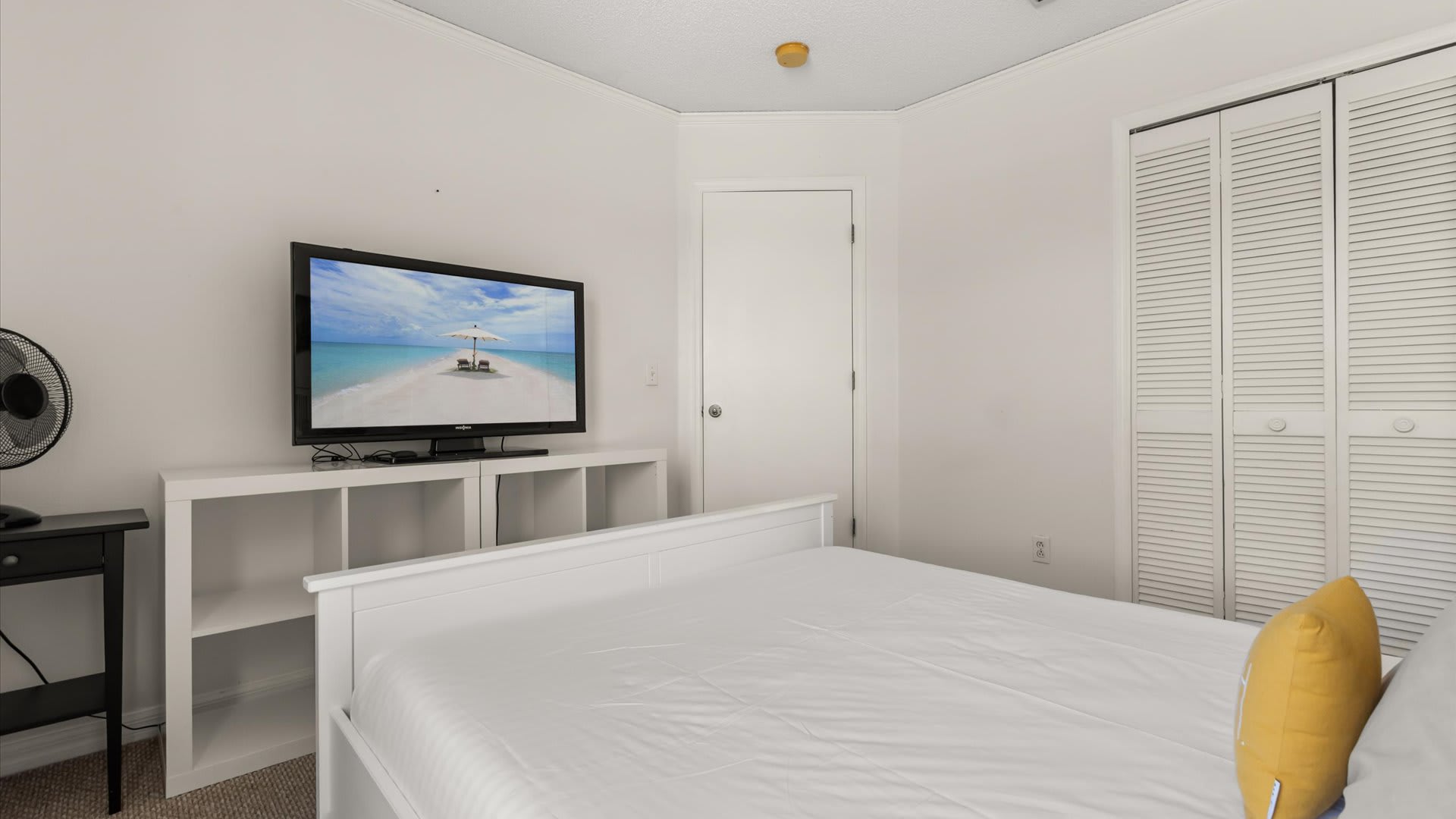 A bedroom with a white wooden bed and a flat-screen TV on a white media console. A closet with louvered bifold doors is visible in the background.