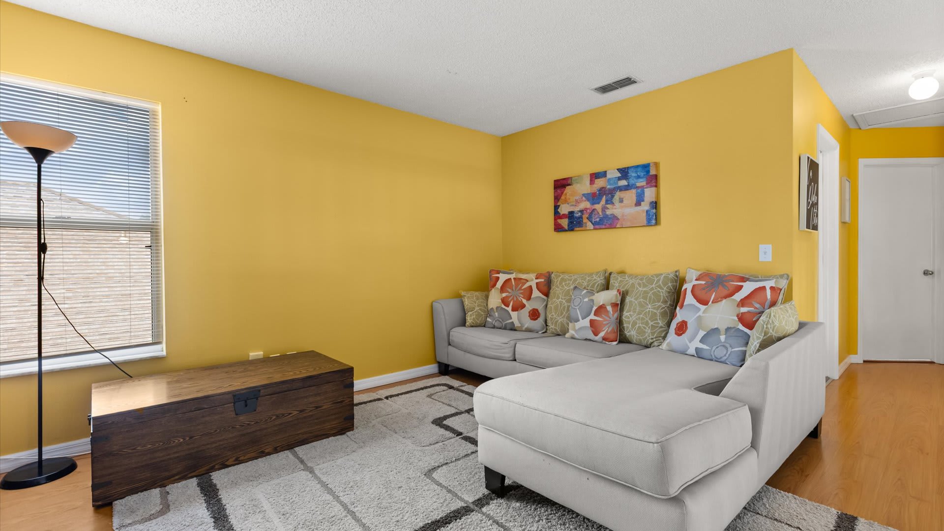 Vibrant living space with bright yellow walls, a light gray sectional sofa adorned with colorful floral pillows, a dark wooden storage chest, and a floor lamp next to a window with blinds.