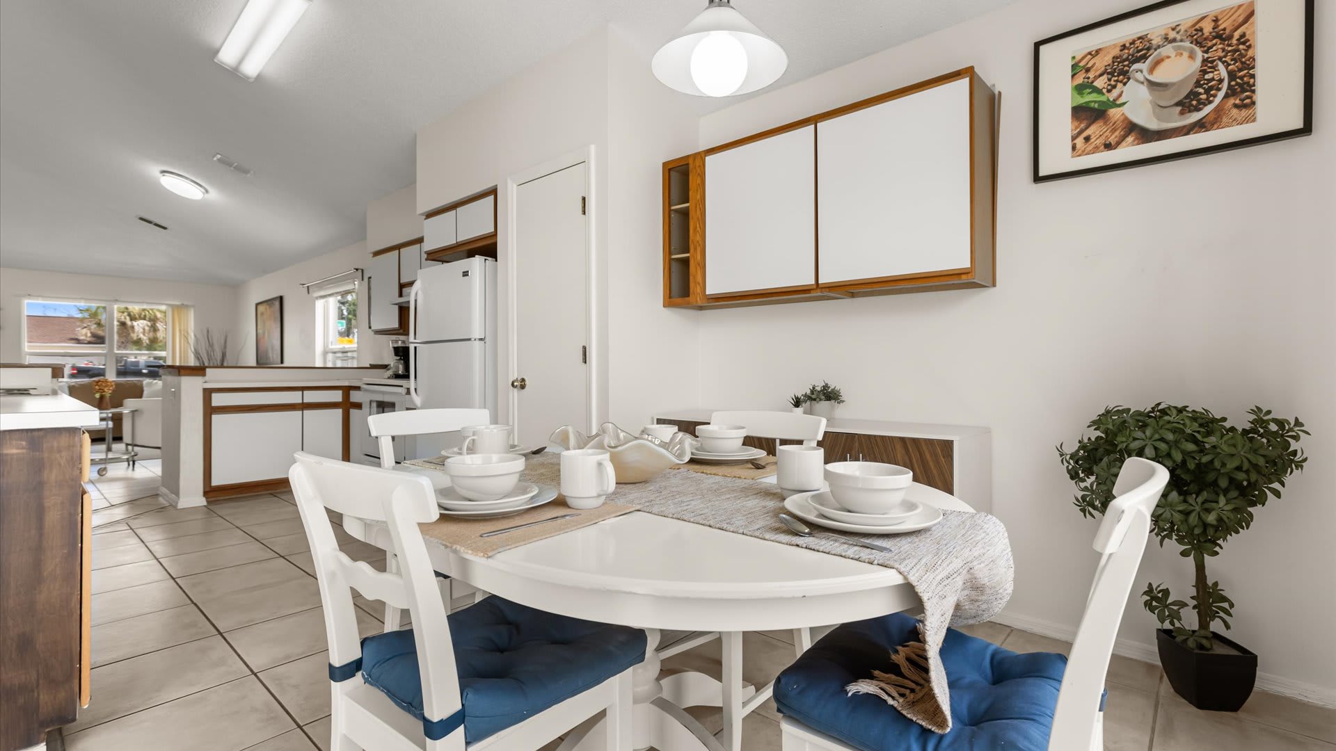 Open-concept dining area with a white round table and blue-cushioned chairs, adjacent to a kitchen with white appliances and wooden accents, featuring a large window and potted plants.