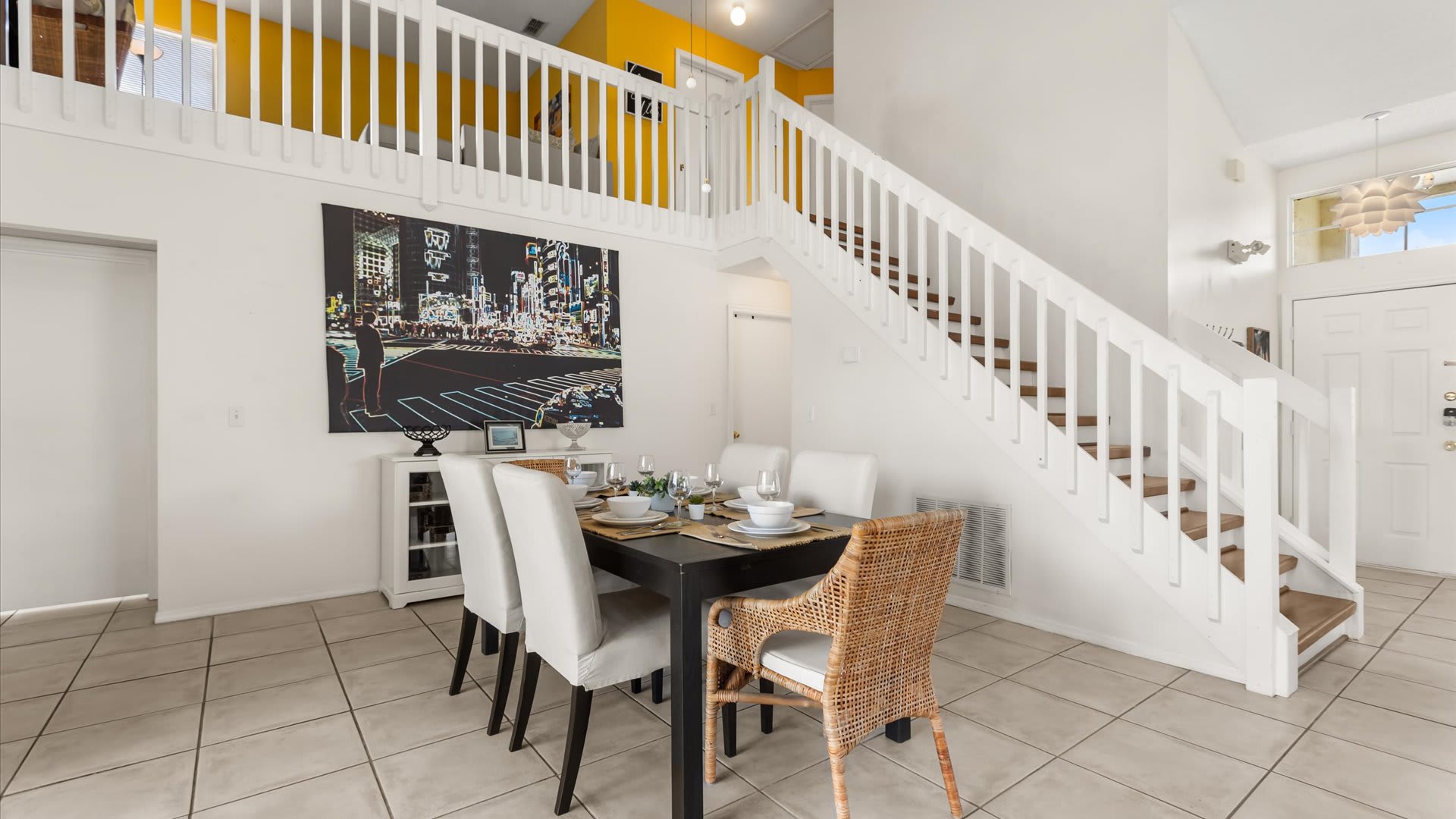 Dining area with a dark table and mixed chairs, positioned beneath a white staircase leading to an upper loft. A large cityscape painting adorns the wall, and the front door is visible.