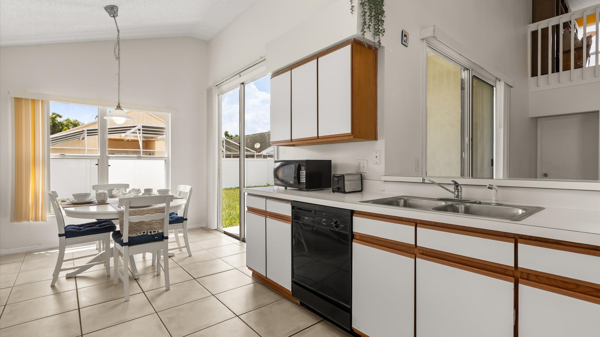 Bright kitchen with white and wood-trimmed cabinets, a black microwave, and a black dishwasher, featuring a small dining area with a white table and chairs next to a large window. Sliding glass doors lead to an outdoor area.