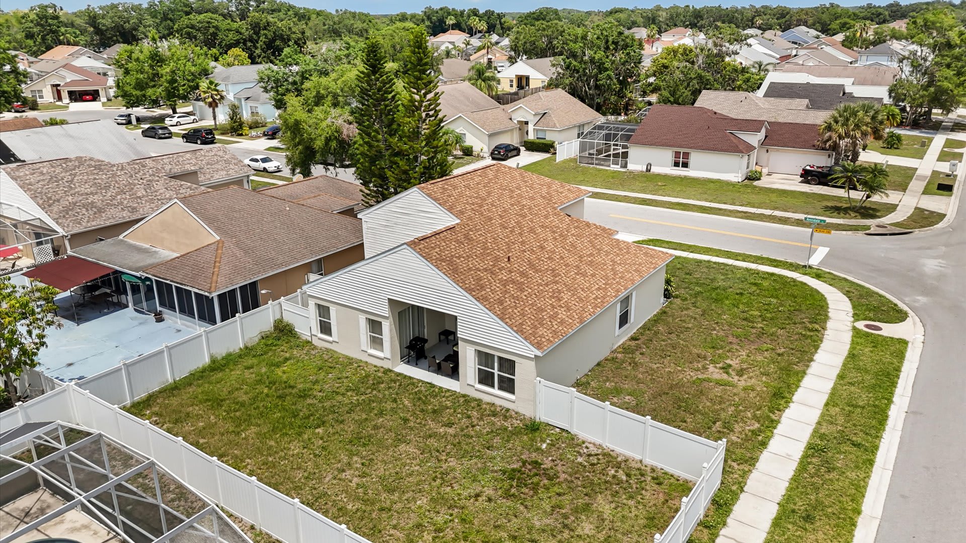 An aerial view of the property showing the light-colored house with a brown shingle roof, a large grassy backyard enclosed by a white fence, and the surrounding residential area with other homes and streets.
