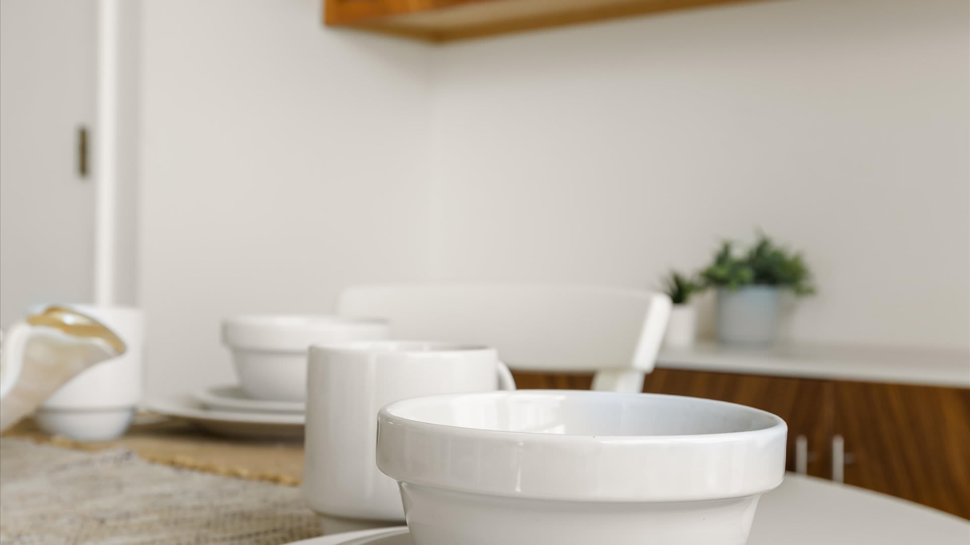 Close-up view of a white dining table set with clean white dinnerware, including bowls and mugs, on a textured placemat, with a blurred background of the dining area.
