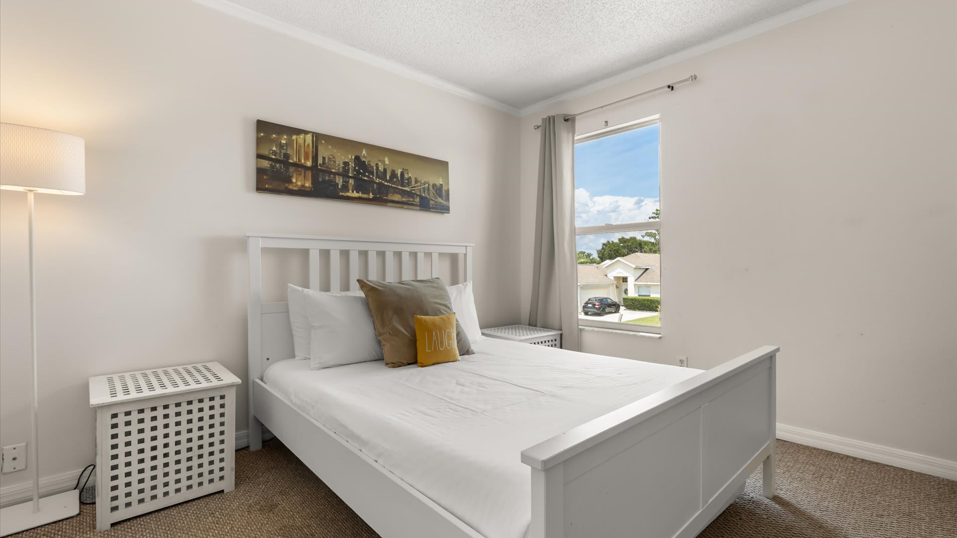 A bright bedroom featuring a white wooden bed frame with white bedding and a yellow accent pillow, flanked by white nightstands. A large window with gray curtains offers a view of the neighborhood.