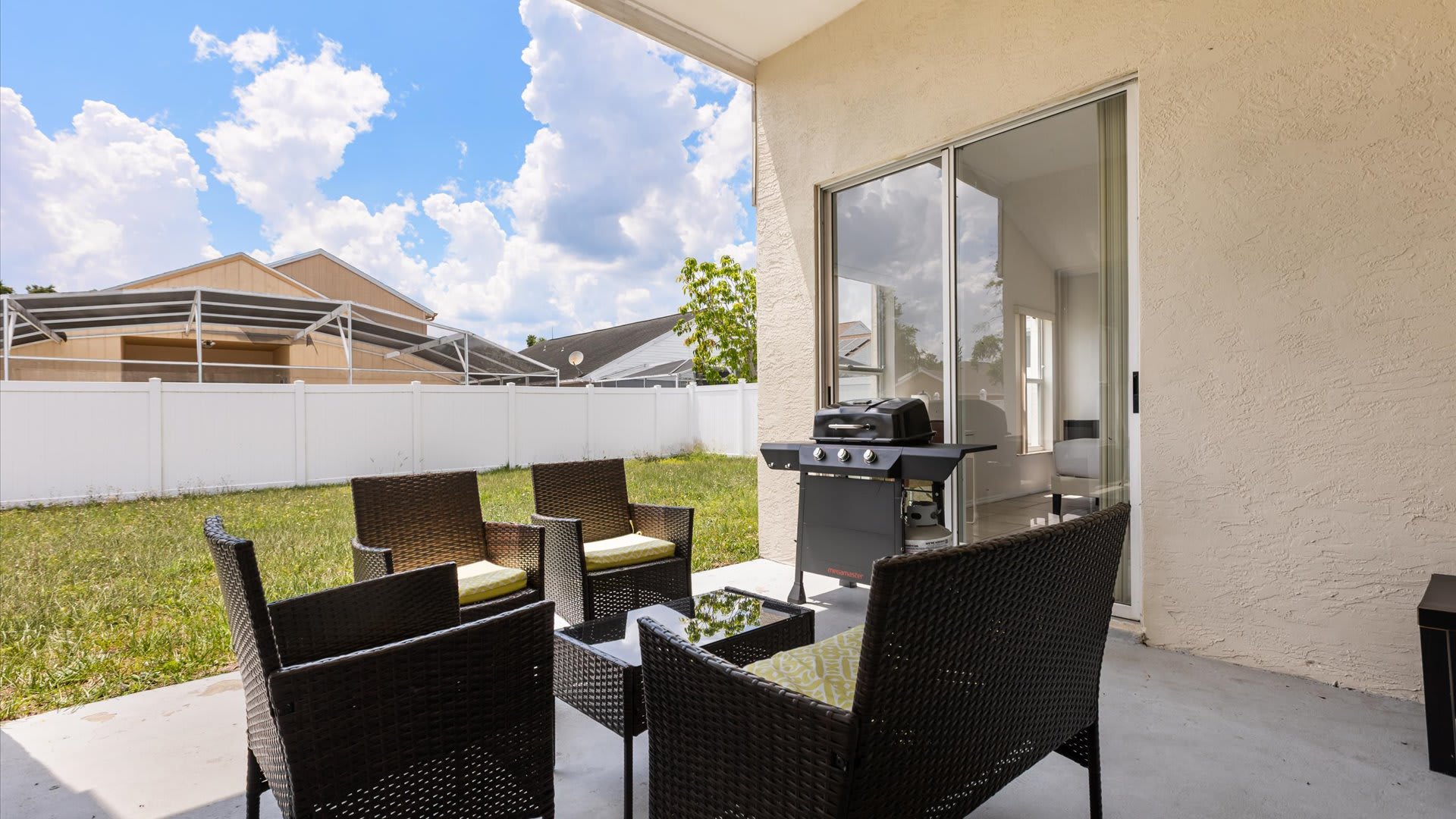 A concrete patio features a dark wicker outdoor seating set with light cushions and a matching coffee table, alongside a black BBQ grill, all under a covered area with a view of a grassy backyard and white fence.