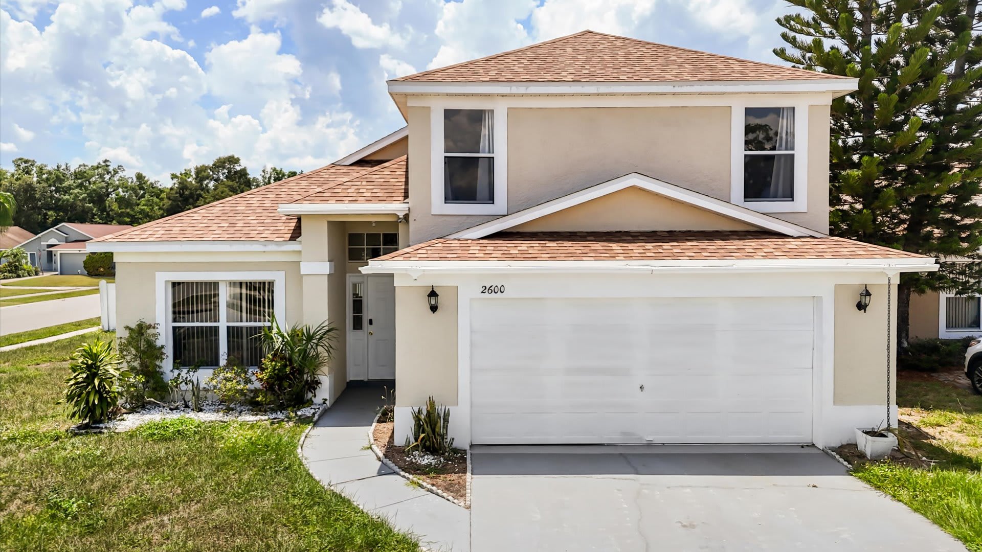 The front exterior of a two-story, light-colored stucco house with a brown shingle roof, featuring a two-car garage, a small landscaped area, and a paved driveway, under a bright blue sky.