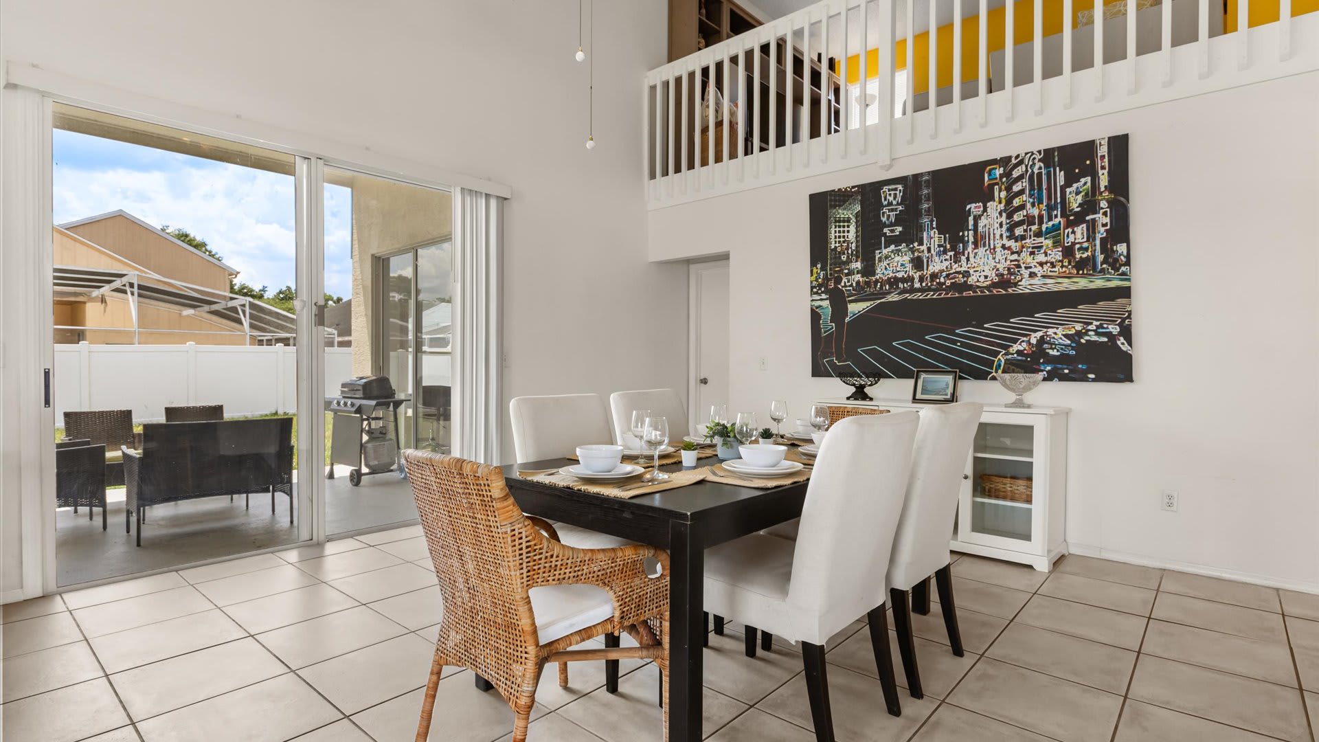 Dining area with a dark rectangular table set for six, featuring white upholstered chairs and one wicker chair. Large sliding glass doors open to an outdoor patio with a grill.