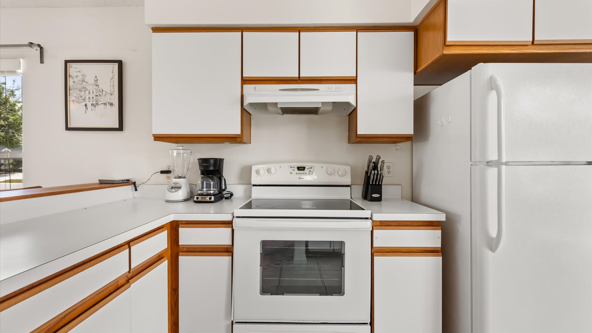 Corner view of a kitchen with white appliances including a stove and refrigerator, white countertops, and white cabinets with wood trim. A coffee maker and blender are visible on the counter.