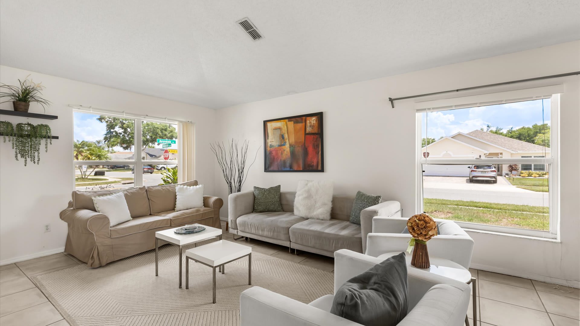 Bright living room with a beige slipcovered sofa and a light gray sofa, white armchairs, and white coffee tables on a geometric rug, featuring large windows with street views and an abstract painting.