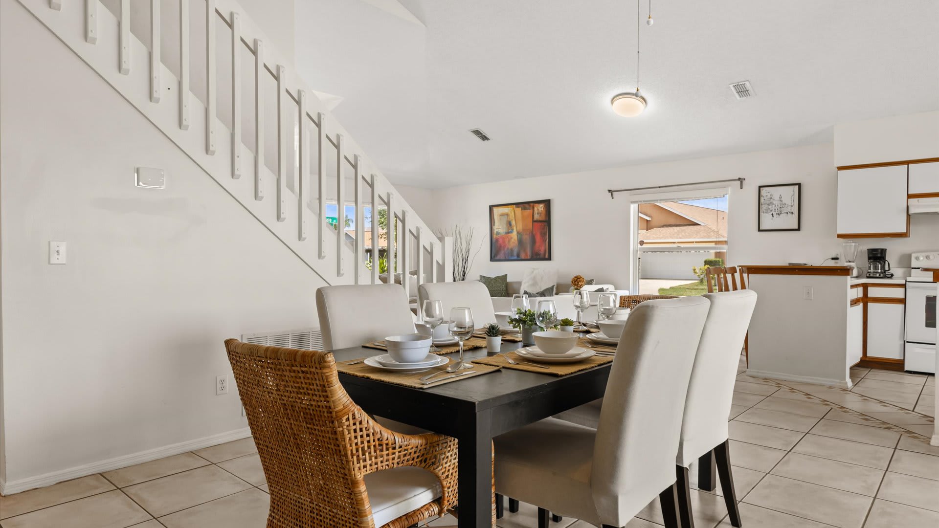 Dining area with a dark table and white chairs, open to the living room and kitchen, featuring a staircase leading upstairs. The room has white walls and tile flooring.