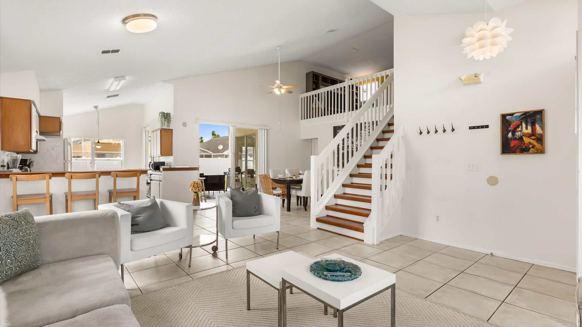 Expansive open-concept living space featuring a light gray sofa, white armchairs, and coffee tables, with a prominent wooden staircase leading to an upper level, and a view into the kitchen and dining area.