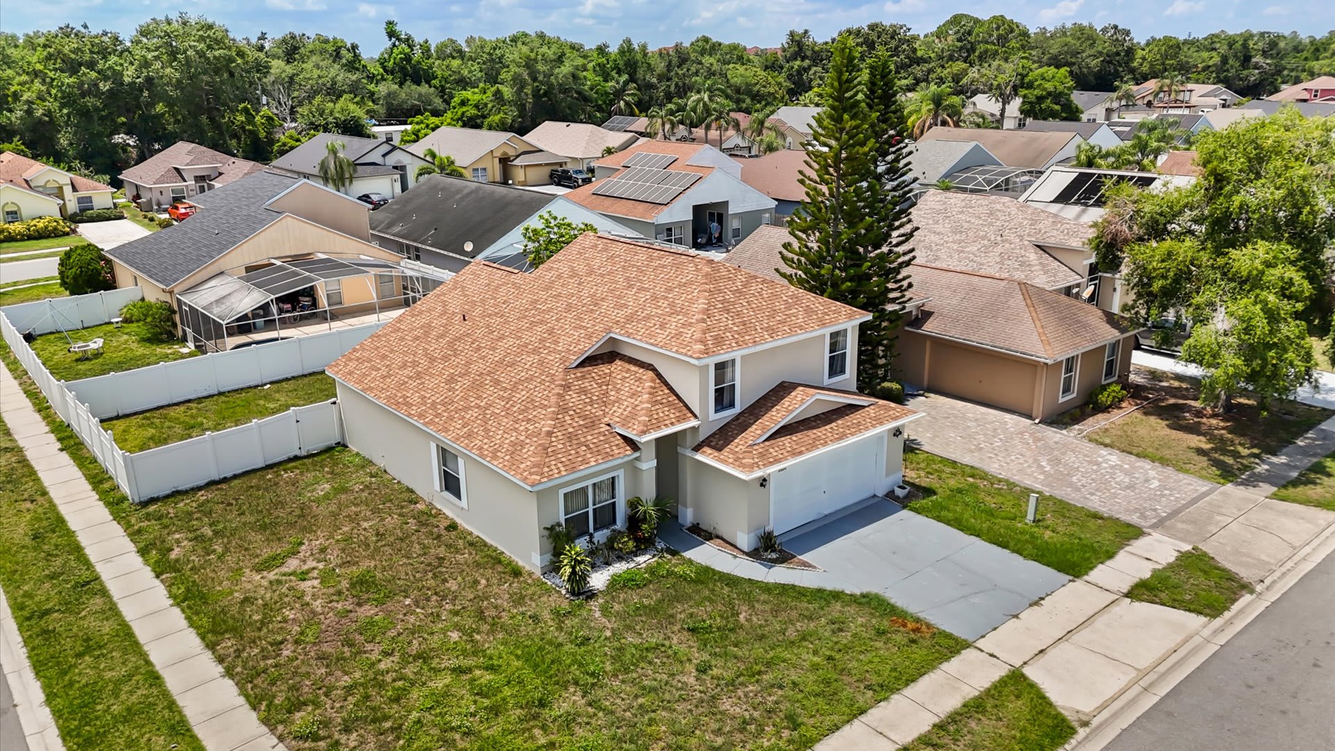 An aerial view showcasing the front and side of a two-story, light-colored house with a brown shingle roof, a two-car garage, and a grassy yard, situated in a suburban neighborhood with a street and other houses.