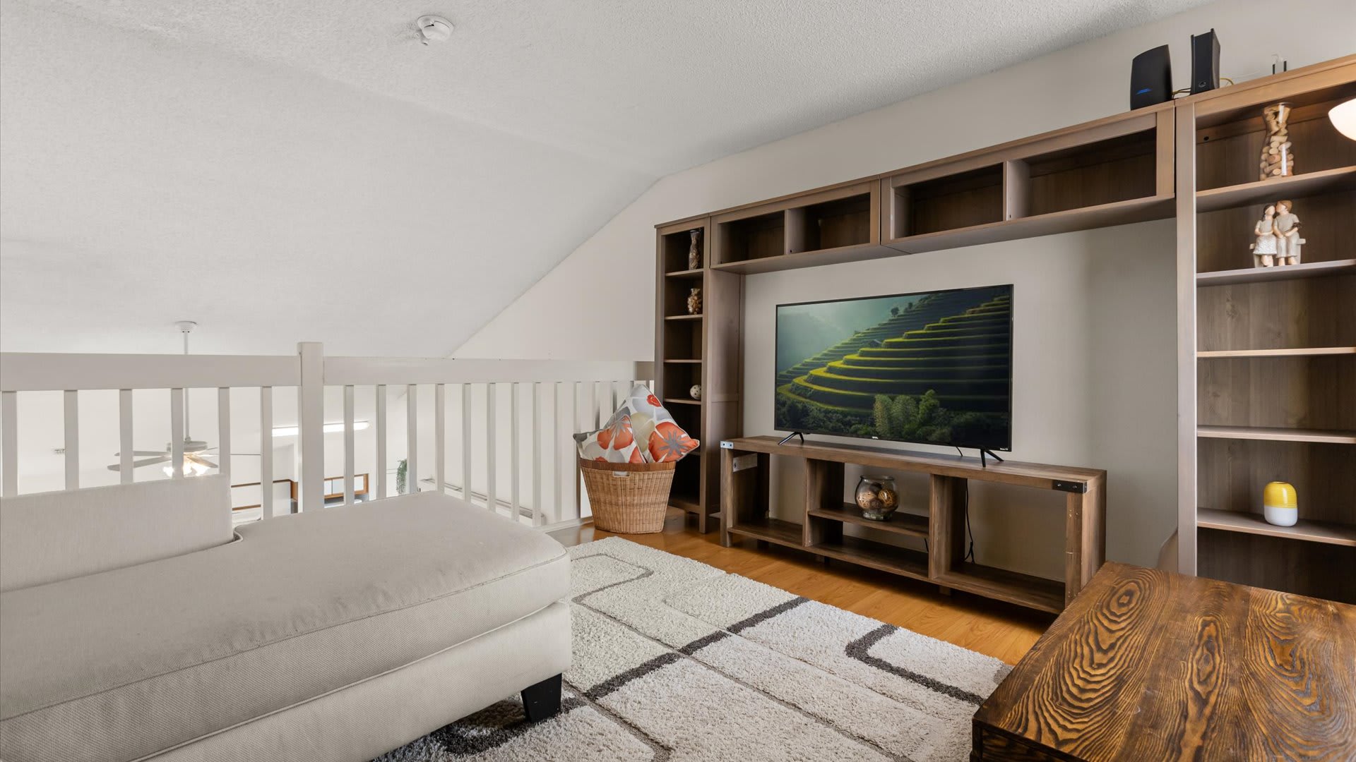 Cozy loft living area with a light-colored sectional sofa, a large flat-screen TV integrated into a wooden shelving unit, and a patterned rug. The space overlooks the main living area below.