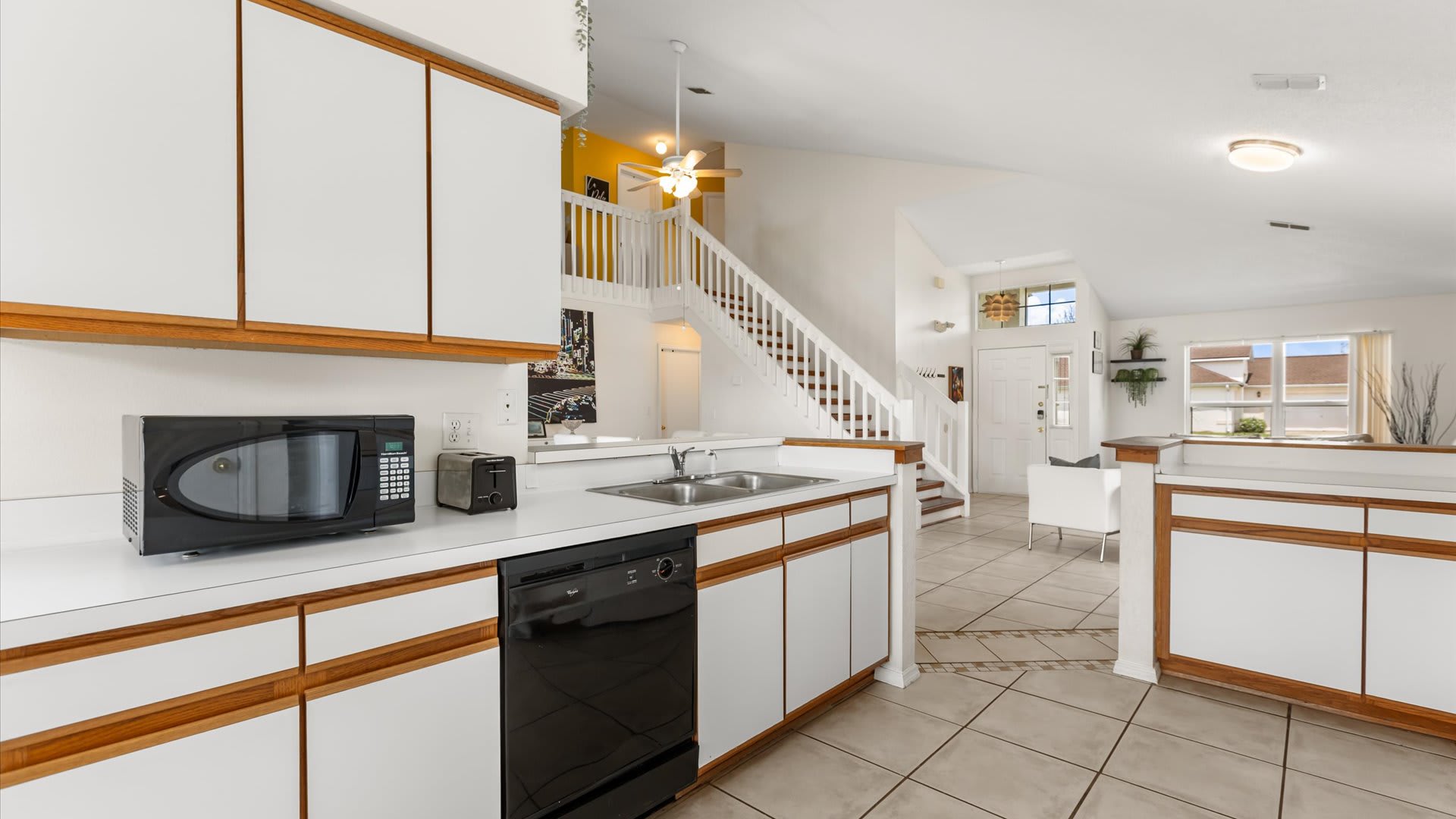 Wide shot of the kitchen showing white and wood-trimmed cabinets, a black microwave, and a black dishwasher, with a view into the living area and staircase. The kitchen features white tile flooring and a double sink.