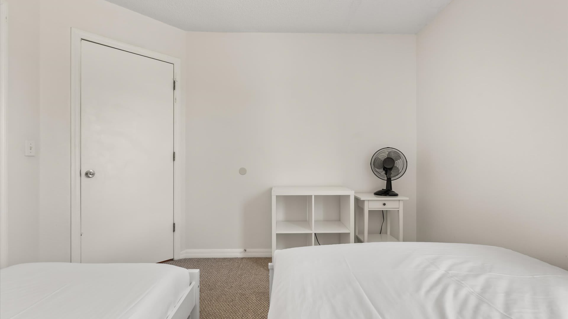 A view of a bedroom showing two white wooden twin beds with white bedding, a white shelving unit, and a black oscillating fan on a small white nightstand. A white door is visible to the left.
