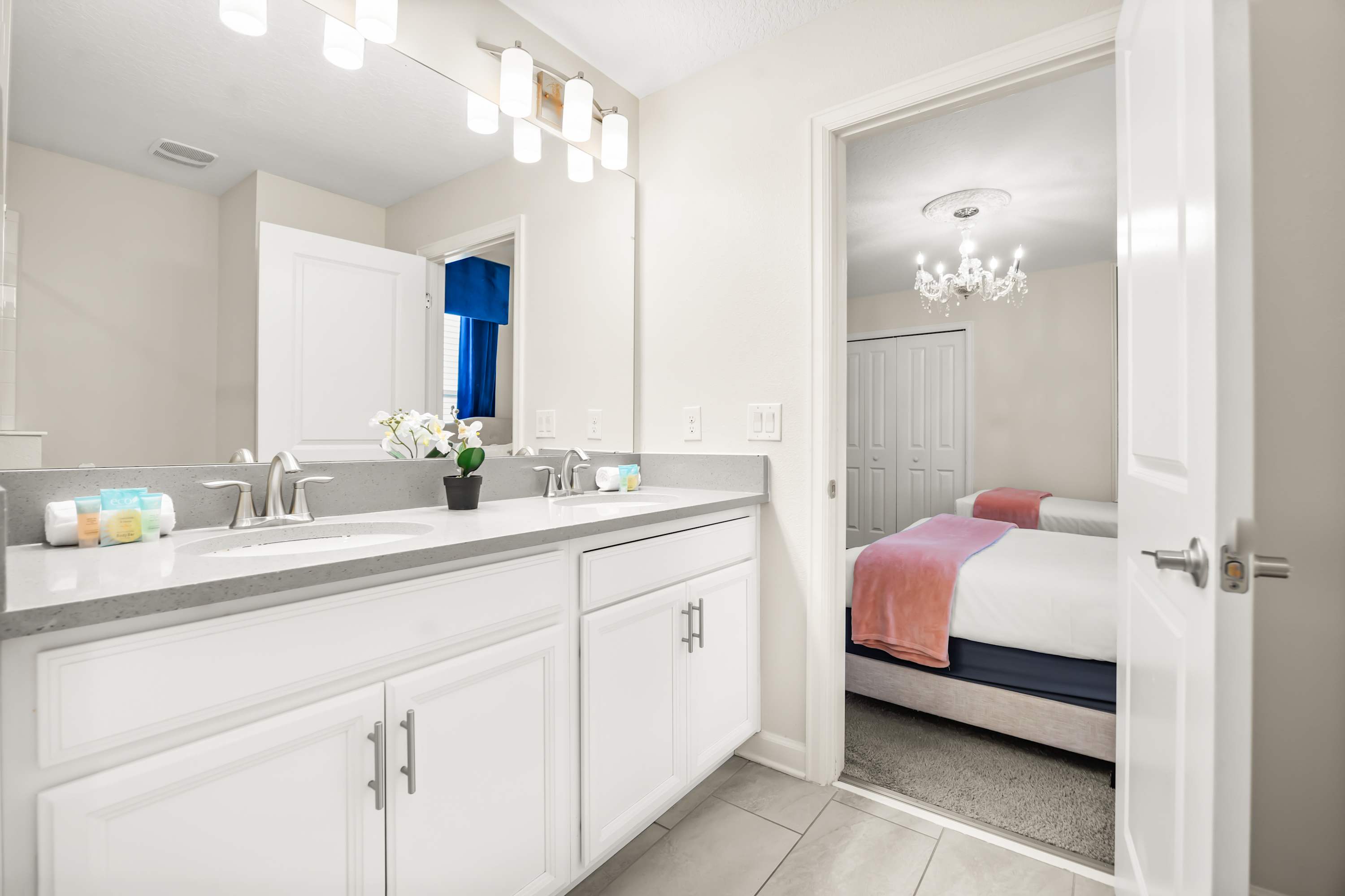 A bright bathroom with a double vanity featuring a light gray countertop, two sinks with brushed nickel faucets, and a large mirror with modern light fixtures above.