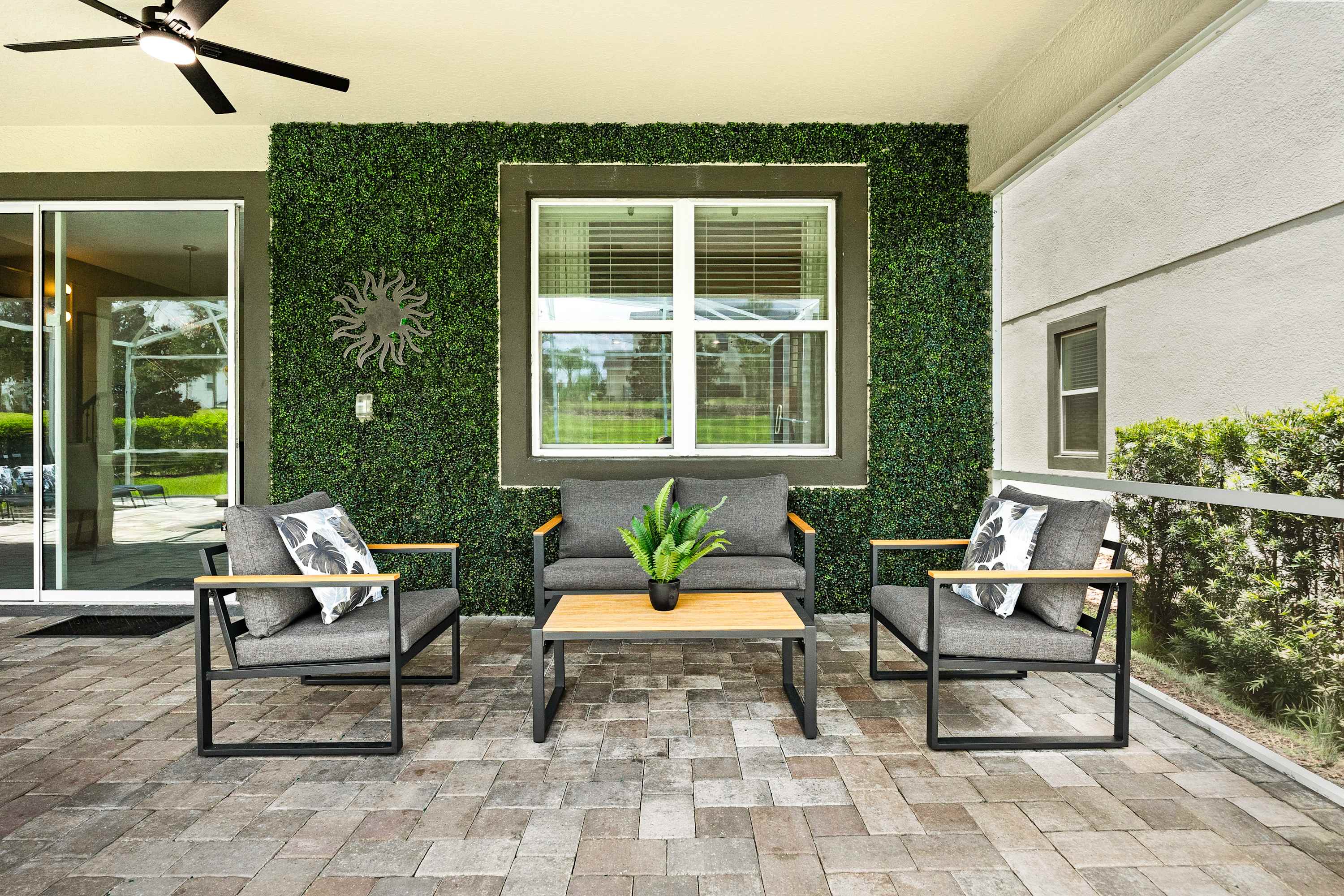 A modern outdoor patio features a gray sofa and two armchairs with black frames, arranged around a light wood coffee table on a stone-tiled floor. A green artificial hedge wall with a sunburst decor piece provides a vibrant backdrop.