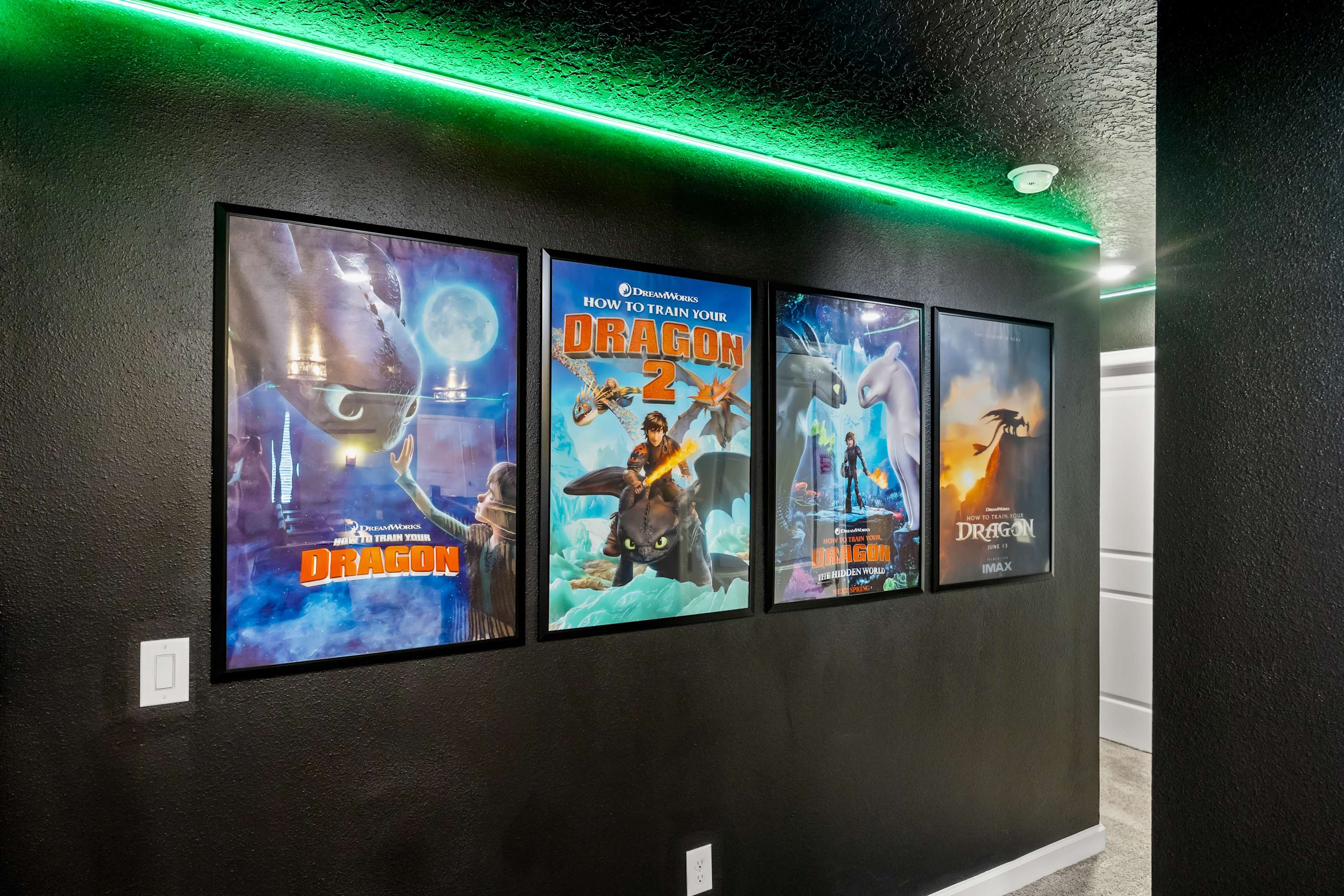 A hallway leading to the media room, decorated with four framed movie posters from "How to Train Your Dragon" against a dark wall. Green LED strip lighting runs along the ceiling, complementing the themed decor.