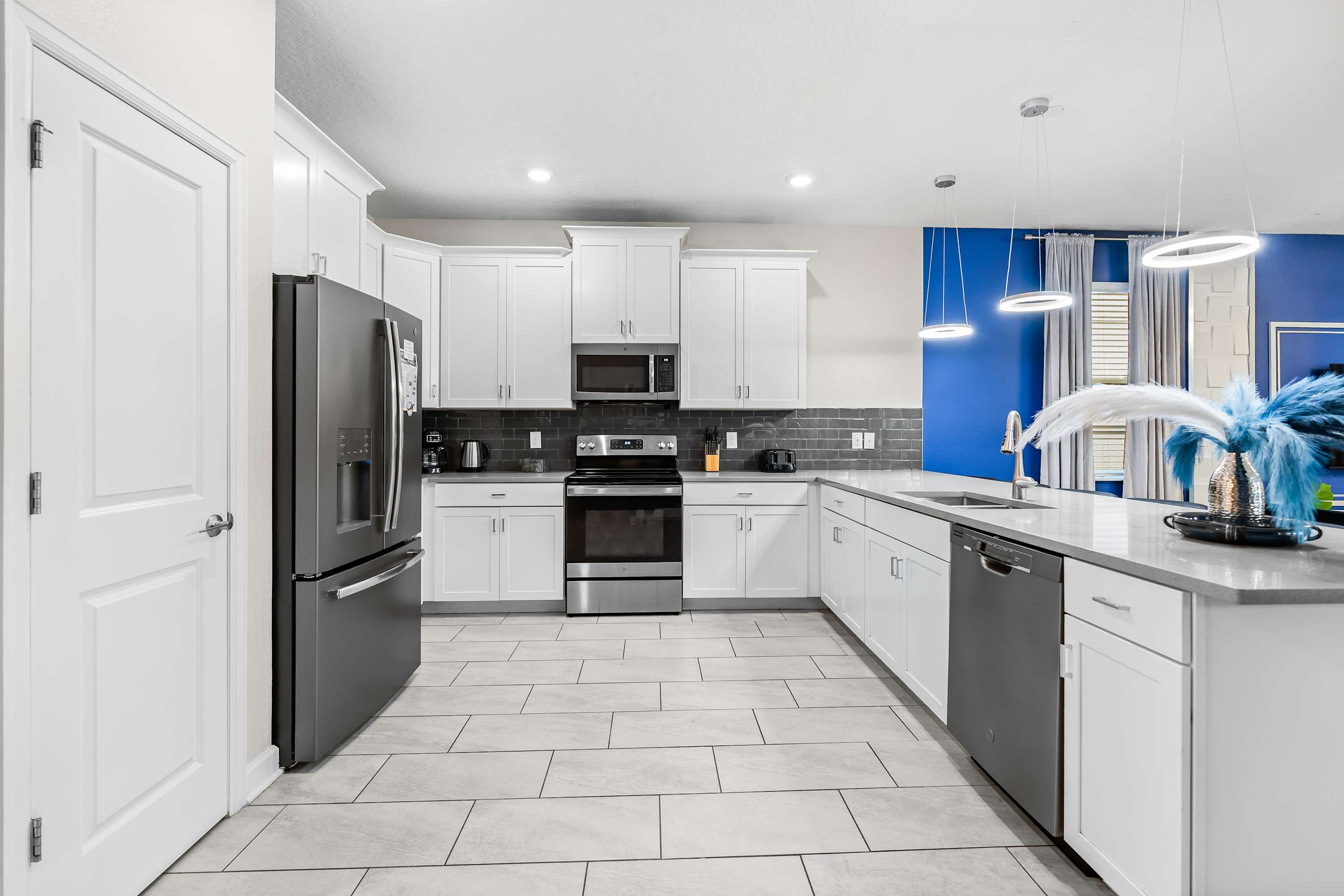 A wide shot of a contemporary kitchen with white shaker cabinets, light gray countertops, and stainless steel appliances including a refrigerator, oven, and microwave. A dark gray subway tile backsplash adds contrast, and the floor is covered in light gray rectangular tiles.