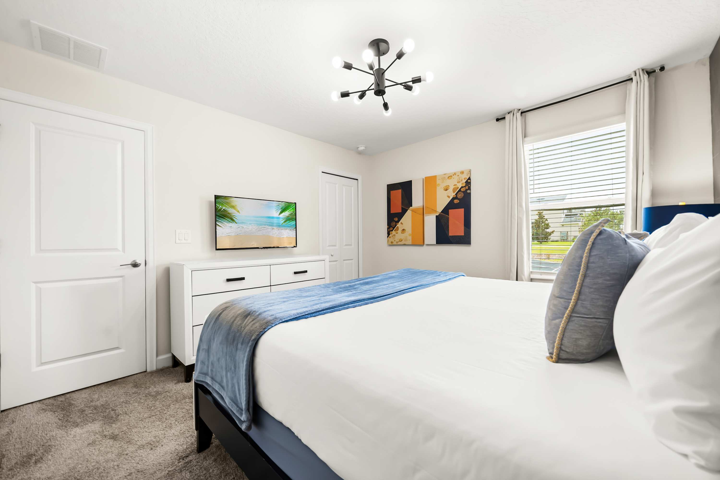 A different perspective of the bedroom, showing a white dresser with a wall-mounted flat-screen TV and a window with white curtains. The room features light-colored walls and a carpeted floor, maintaining a contemporary feel.