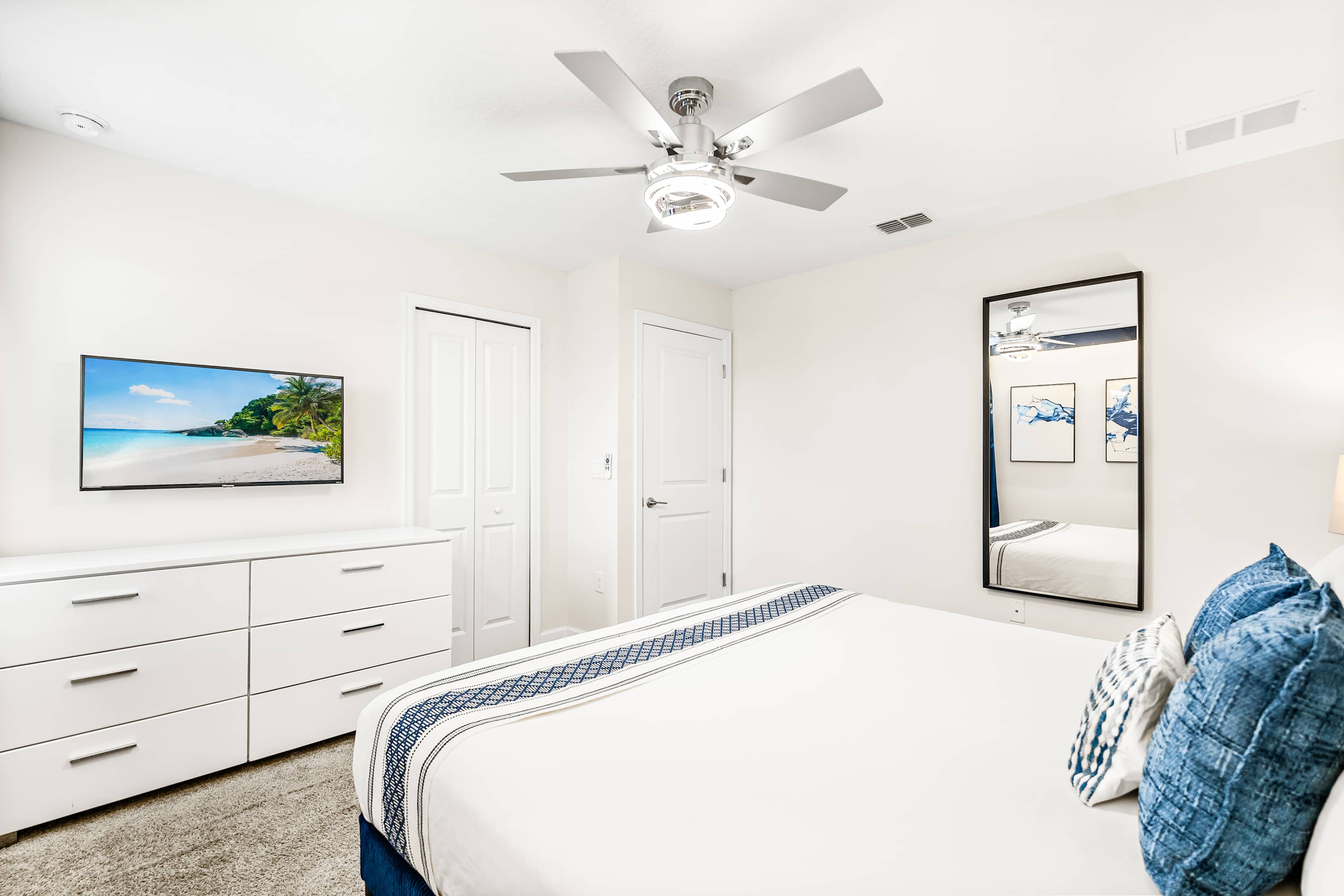 Another view of the modern blue-themed bedroom, showcasing a white dresser with a flat-screen TV, a full-length mirror, and white closet doors, maintaining the clean and contemporary aesthetic.