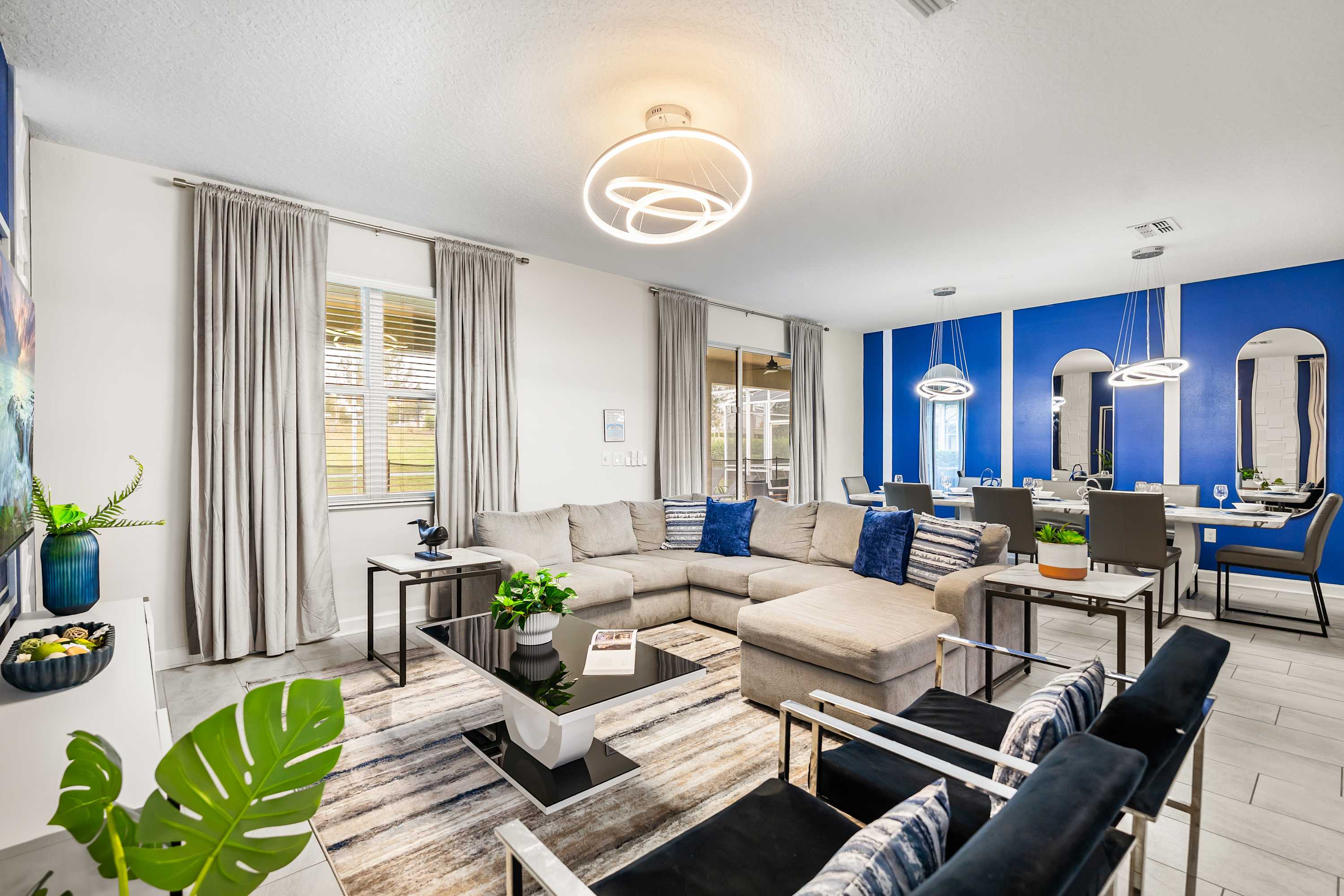 Modern living room with a large L-shaped sectional sofa, two armchairs, a coffee table, and a striped area rug. A dining area with a long table and blue accent wall is visible in the background.