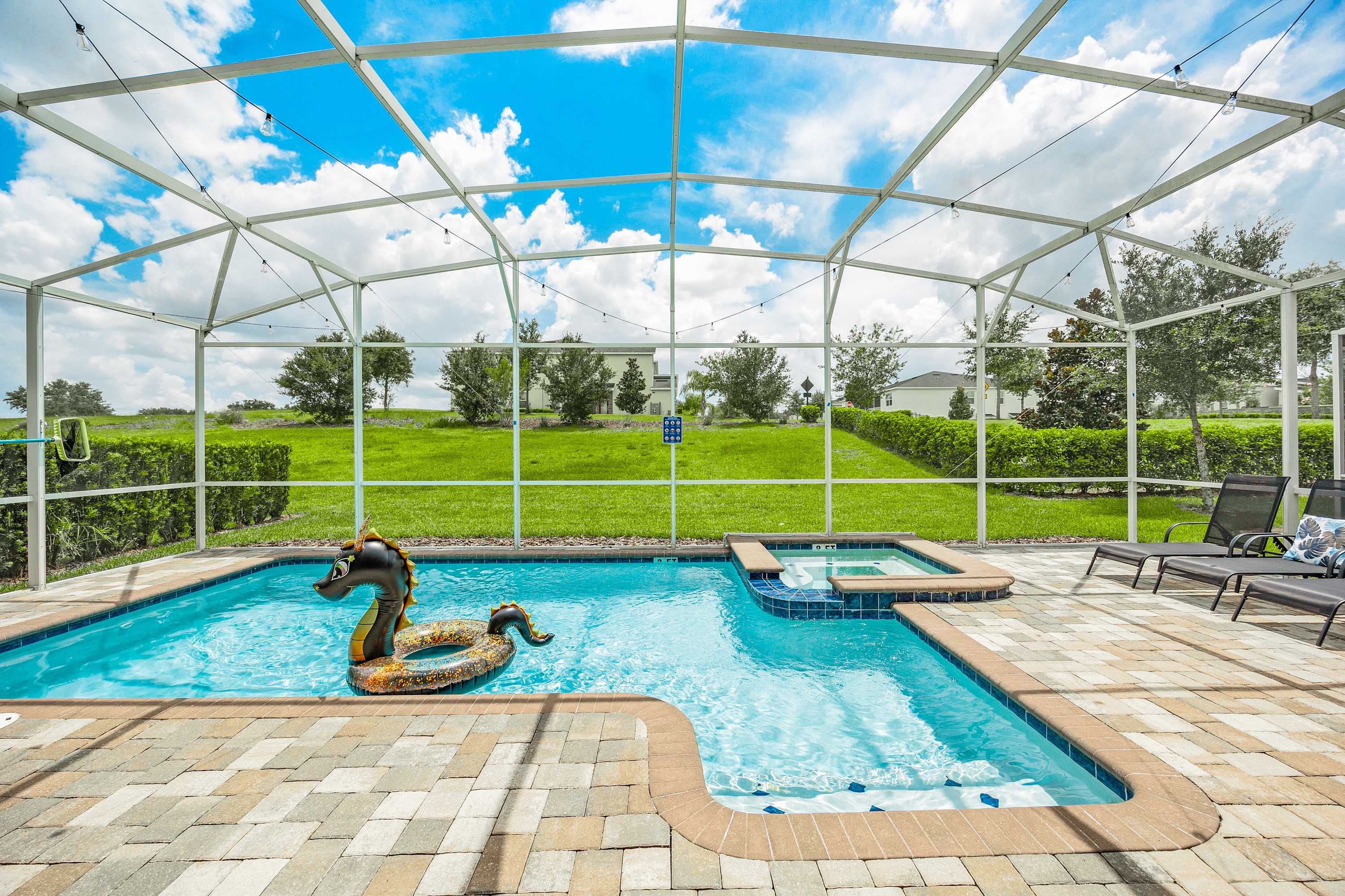 Bright outdoor pool area within a screened enclosure, featuring a rectangular swimming pool with a floating inflatable, an adjacent hot tub, and lounge chairs on a paver deck.