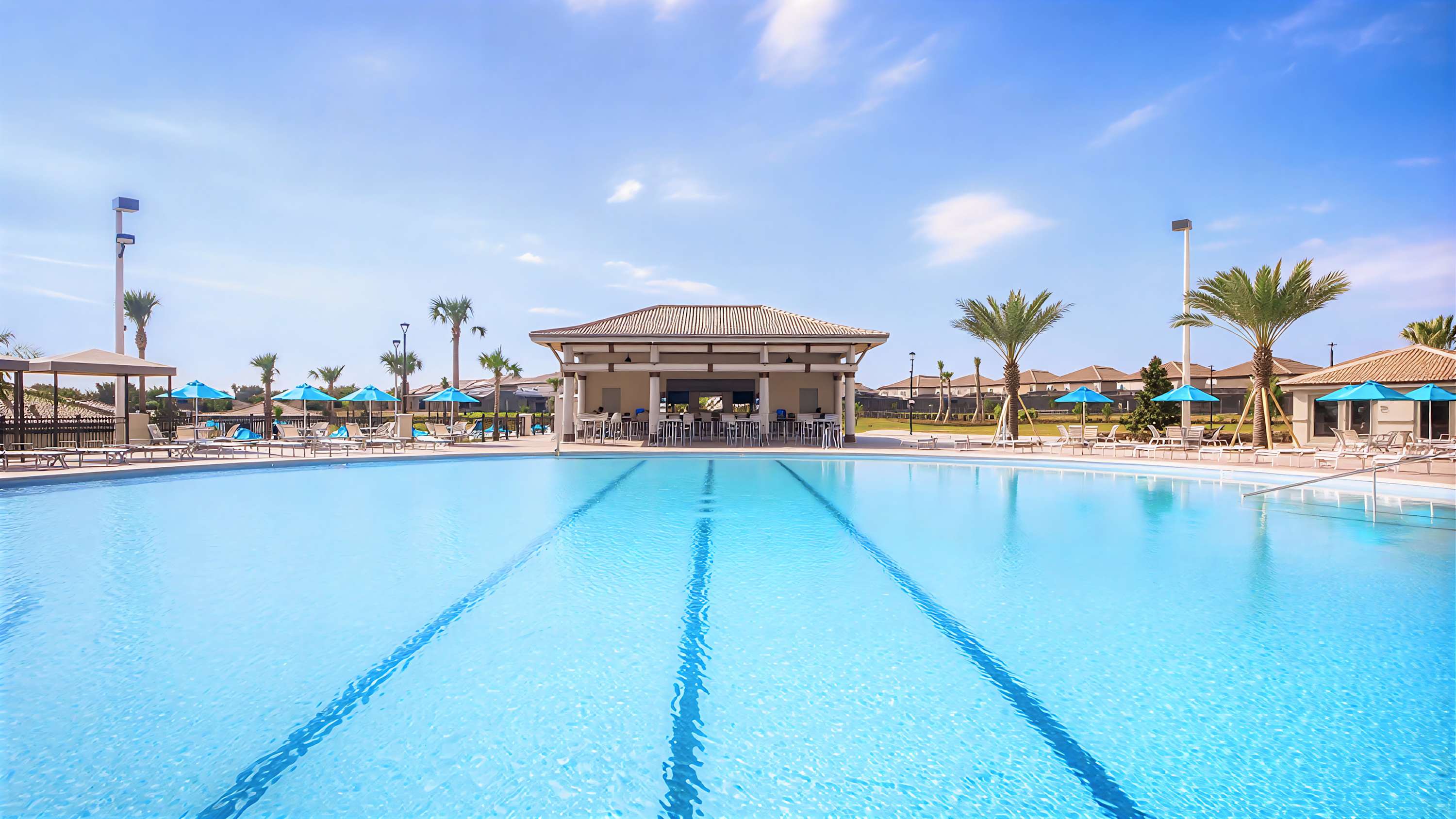 Expansive communal swimming pool with lane markers, surrounded by a spacious deck with lounge chairs and umbrellas, and a covered bar area in the background under a clear blue sky.