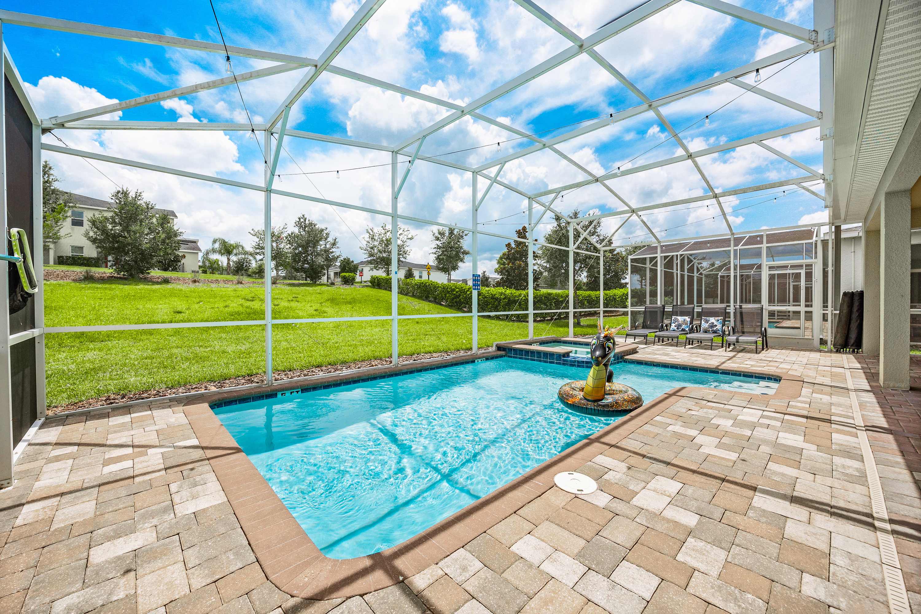 Screened outdoor pool area with a rectangular swimming pool, an integrated hot tub, and several lounge chairs on a paved deck, surrounded by a grassy yard and distant houses under a blue sky.