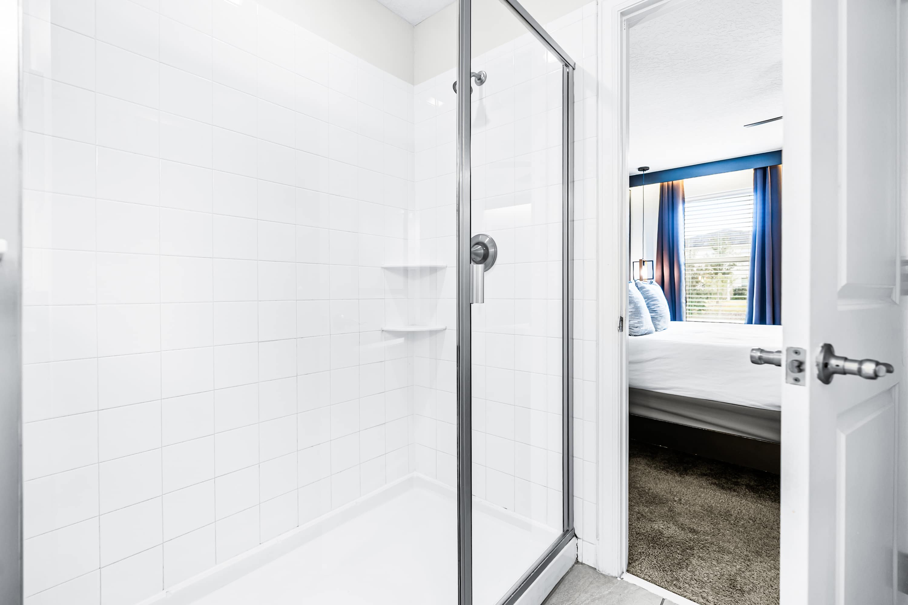 A clean, white-tiled shower stall with a glass enclosure and a built-in shelf, offering a partial view into a bedroom with blue curtains through the open door.