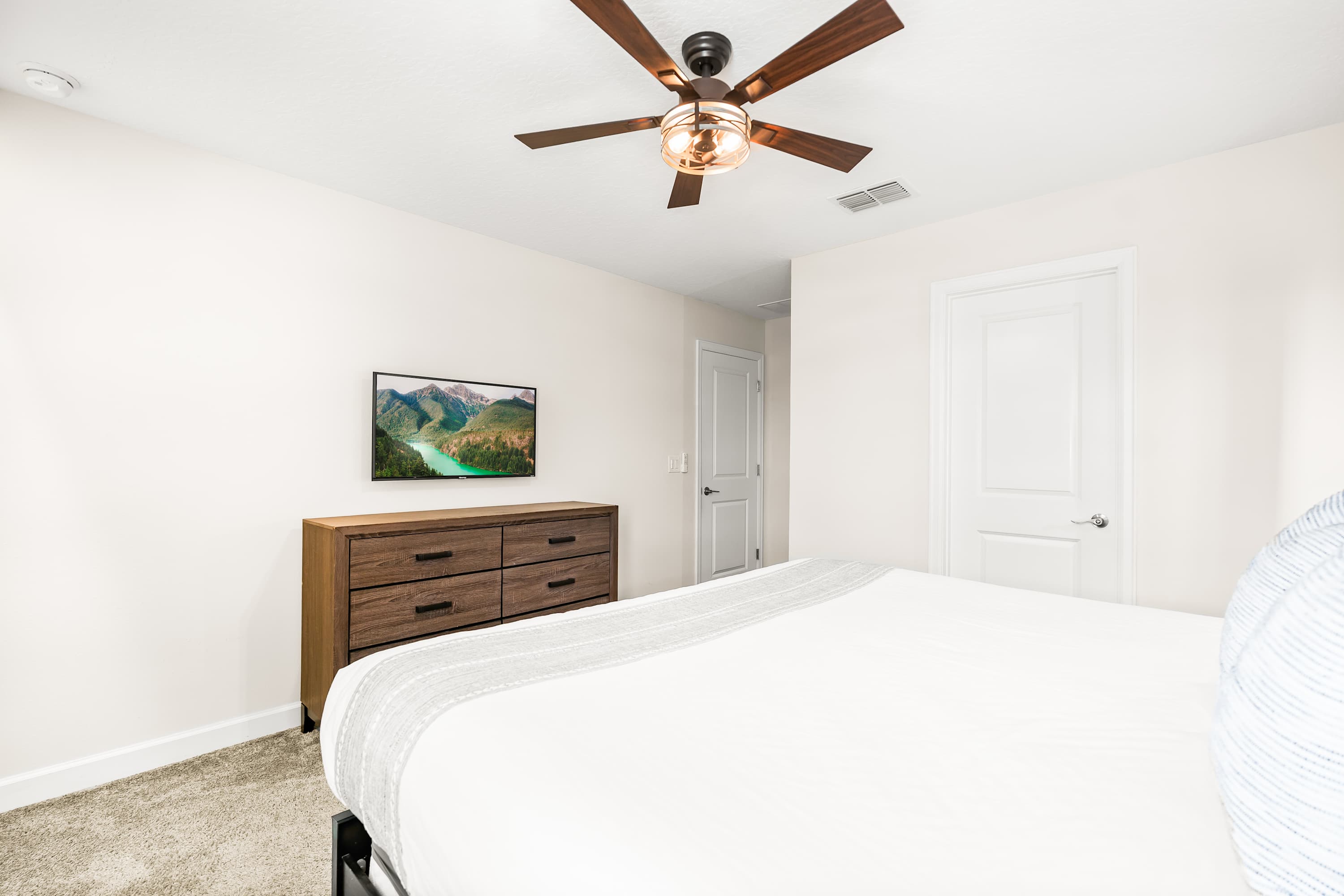 A different angle of the wooden-themed bedroom, highlighting a wooden dresser with a flat-screen TV, white closet doors, and a light-colored throw blanket on the bed.
