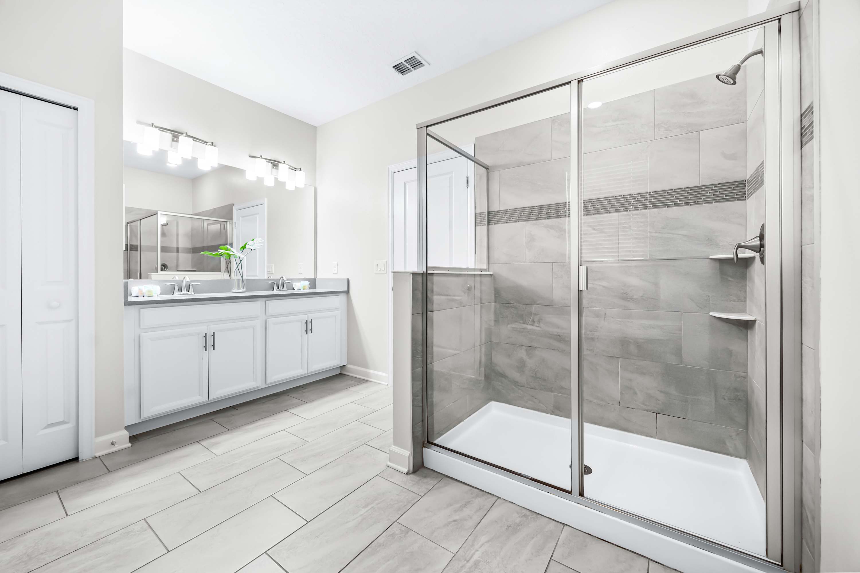 Another view of the bathroom, showcasing a large walk-in shower with glass enclosure and gray tiled walls, alongside a double vanity with white cabinets and a large mirror. The room maintains a clean, modern aesthetic with light gray floor tiles.