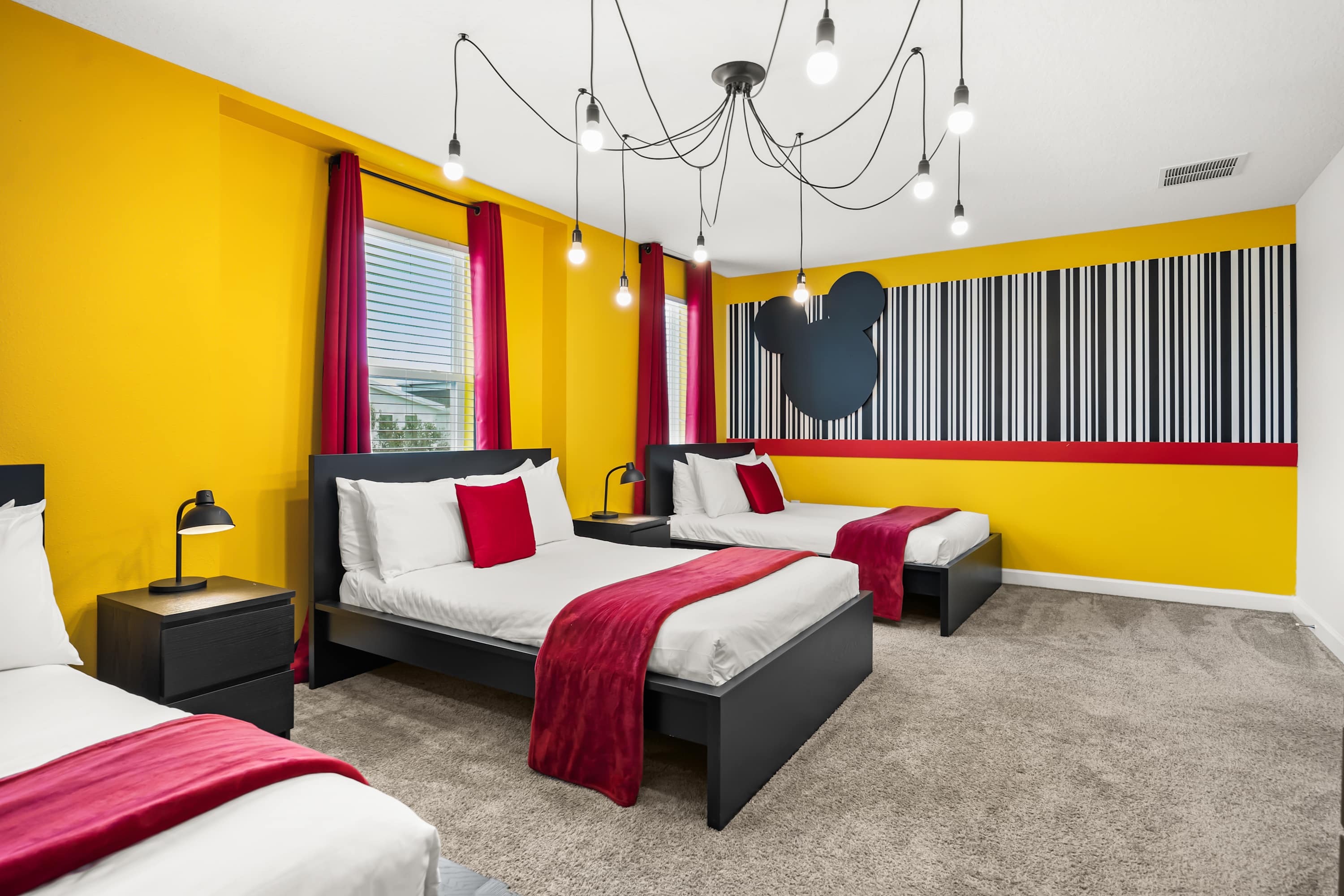 A wider view of the "Minnie Mouse" themed bedroom, showcasing three full beds, black nightstands, and a unique multi-bulb ceiling light fixture. The vibrant yellow walls and Minnie Mouse decor create a playful atmosphere.
