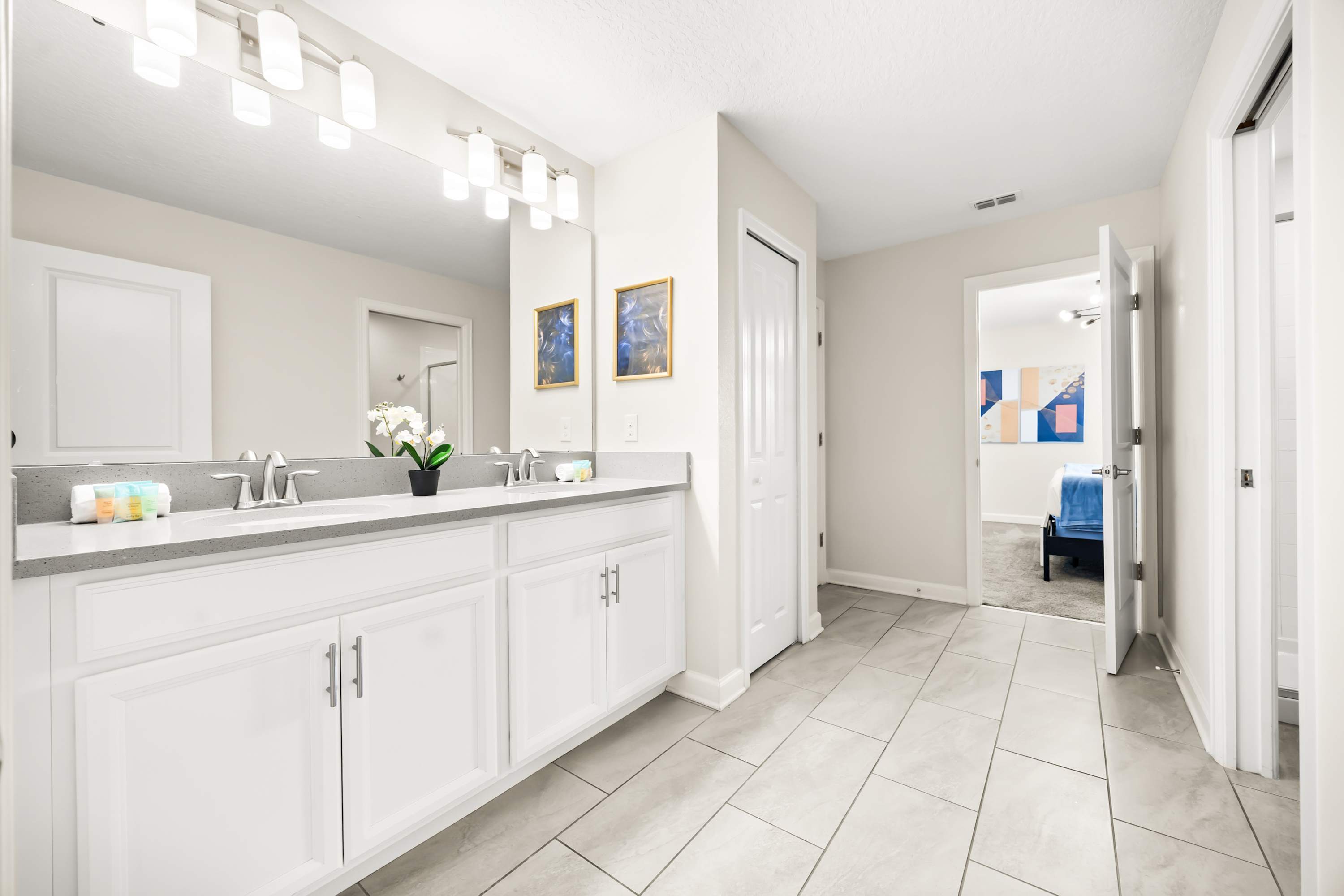 A bright bathroom featuring a double vanity with light gray countertops, white shaker cabinets, and two large mirrors. Light gray tiled floors extend into the adjacent hallway, leading to a bedroom with a blue bed.