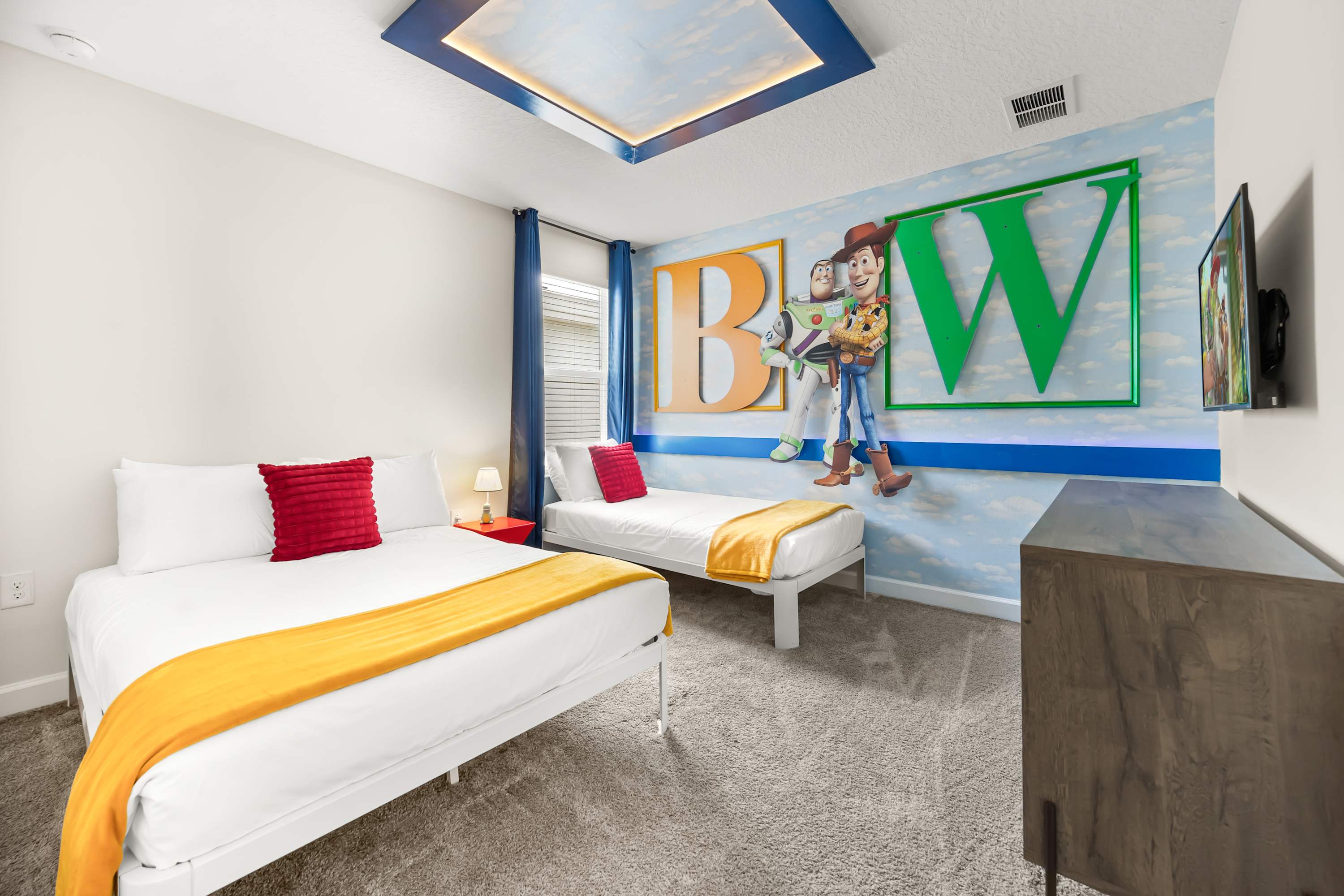 A vibrant "Toy Story" themed bedroom featuring two single beds with white frames, red pillows, and yellow throws. A large mural of Buzz Lightyear and Woody with "BW" letters adorns the wall, complemented by a unique ceiling light fixture.