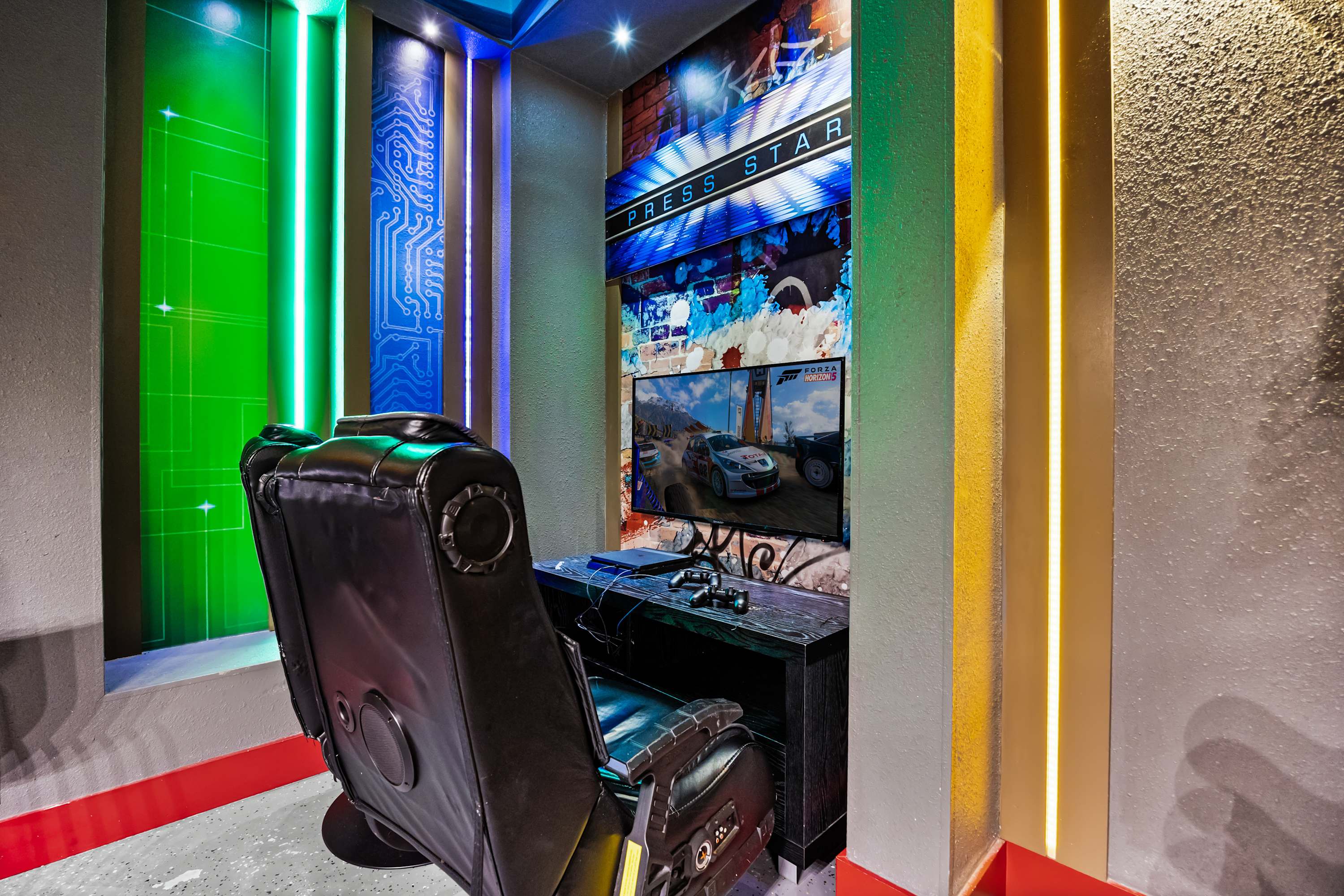 A dedicated gaming nook with a large black gaming chair facing a TV displaying a racing game, set against a vibrant "PRESS START" graphic wall. Green and blue LED strip lights illuminate the textured walls, enhancing the immersive experience.