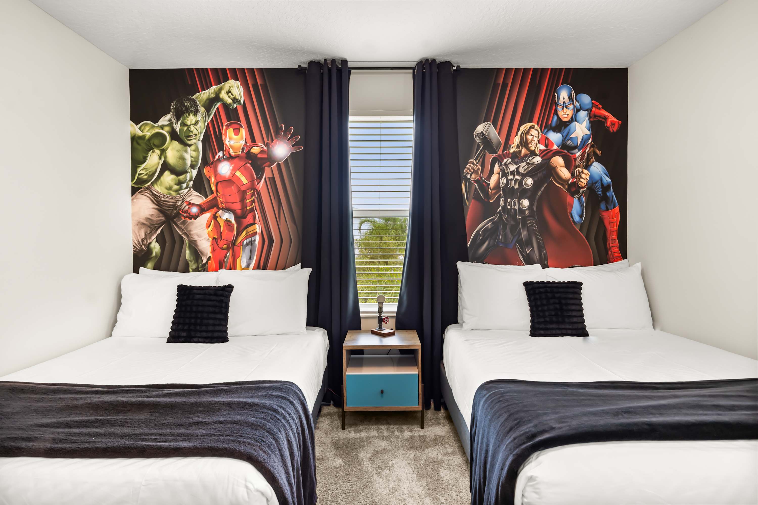 A close-up view of the "Avengers" themed bedroom, showing two full beds separated by a modern nightstand with a blue drawer. The vibrant superhero murals are prominent, and a window with blue blinds is visible between the beds.