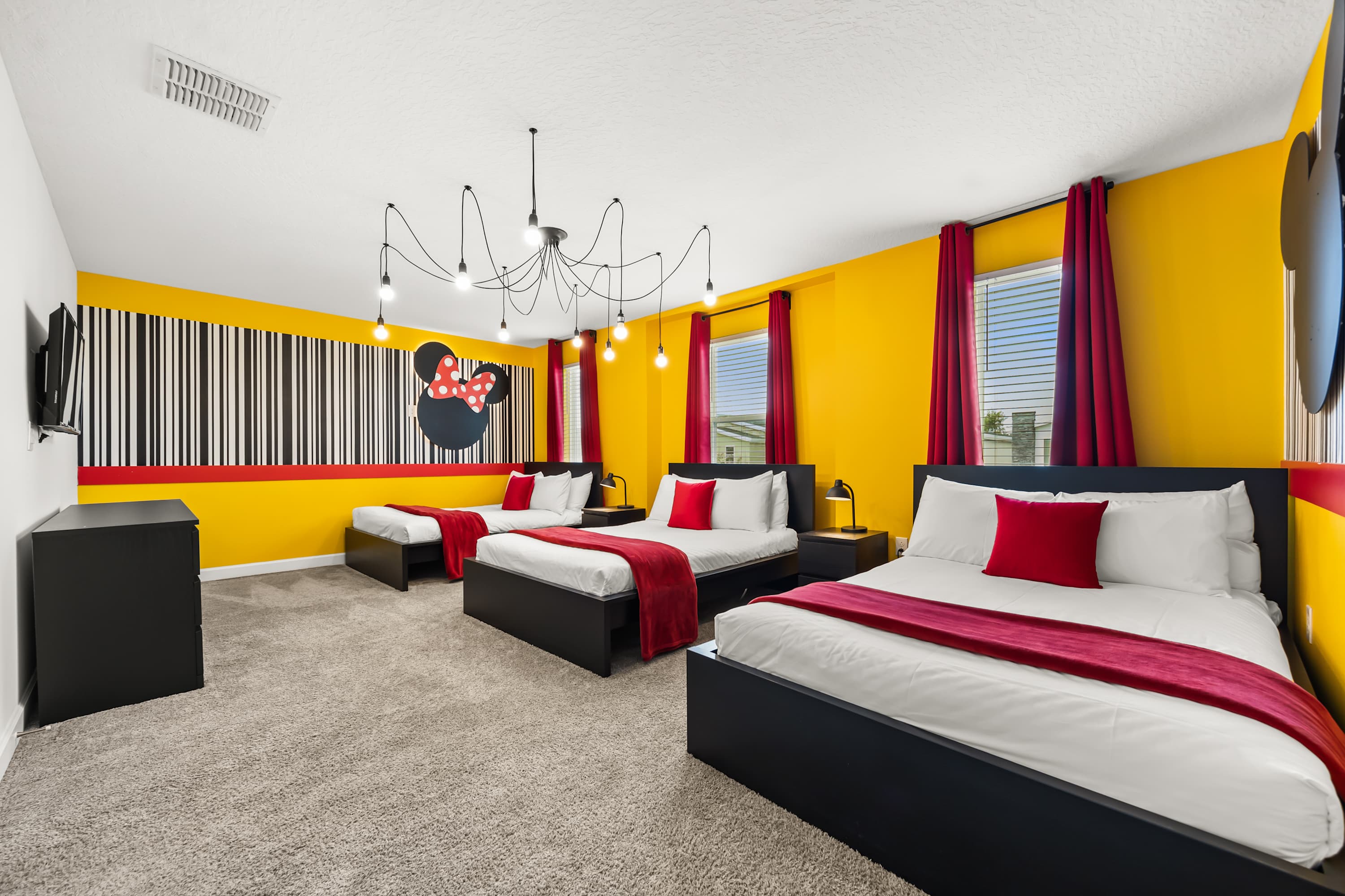 A bright "Minnie Mouse" themed bedroom with three full-sized beds, black frames, red throws, and red pillows. The walls are painted yellow with a black and white striped accent wall featuring a large Minnie Mouse silhouette.