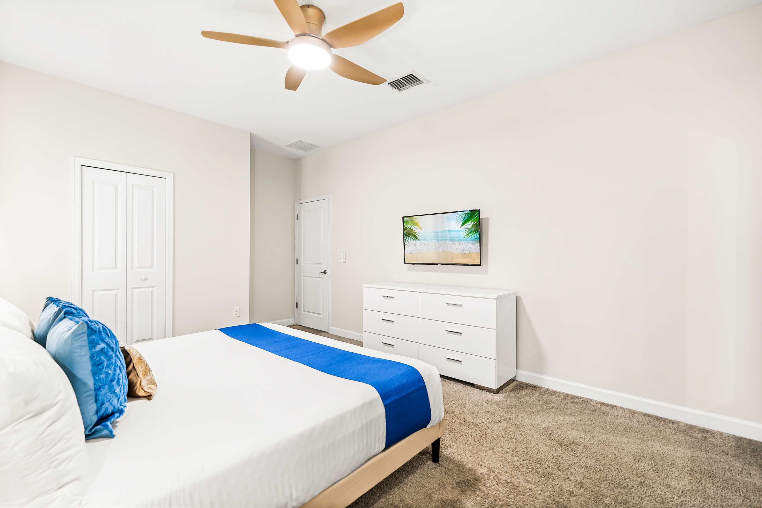 A view of the bedroom from a different angle, showing a white dresser with a wall-mounted flat-screen TV displaying a beach scene. The room has light-colored walls, a ceiling fan, and a carpeted floor.
