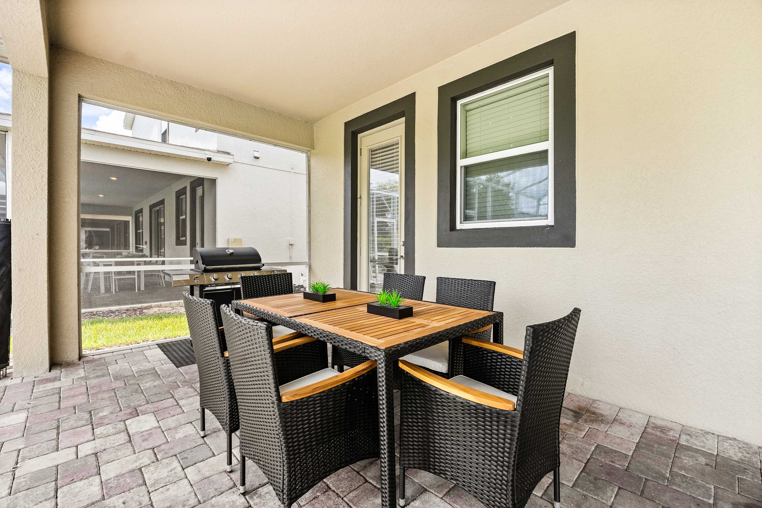 Covered outdoor patio featuring a rectangular wooden dining table with six wicker chairs and a BBQ grill, situated on a paved surface next to the house.